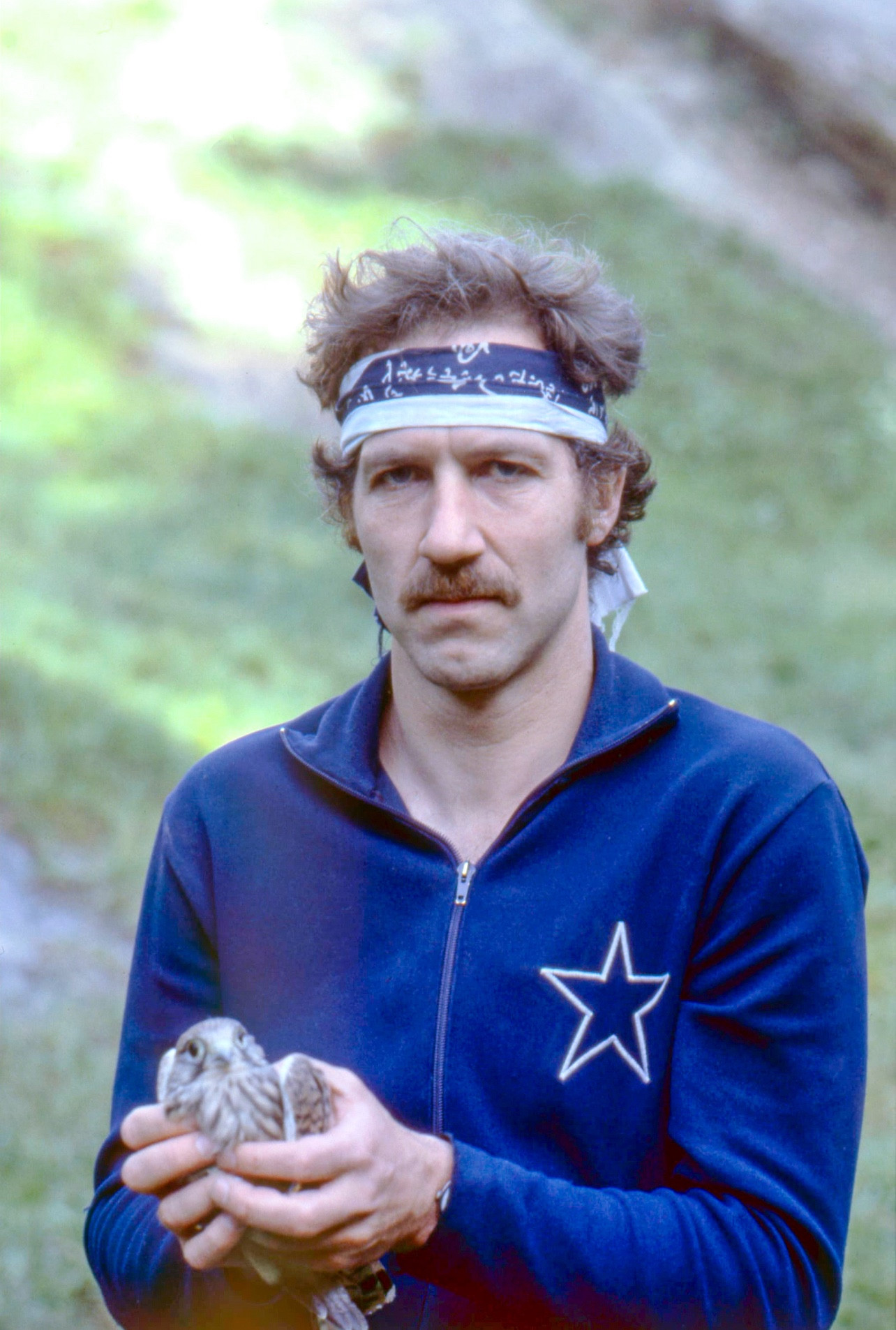 Werner Herzog standing in a field in front of a hill, wearing a blue track suit with a star emblem on the front, and a blue headband. He’s holding a small bird in his hands.