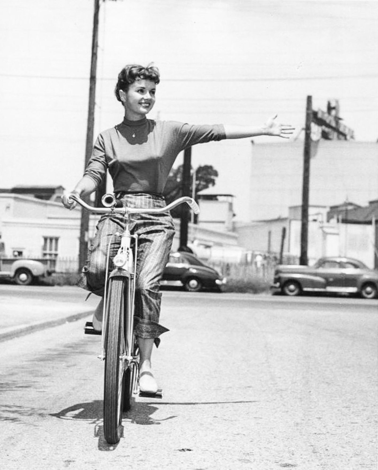 Debbie in a tight long sleeved shirt and striped pants, riding on a bicycle on an empty street. She’s extending her left arm in a signaling motion. There are some houses, telephone poles and parked cars behind her. 