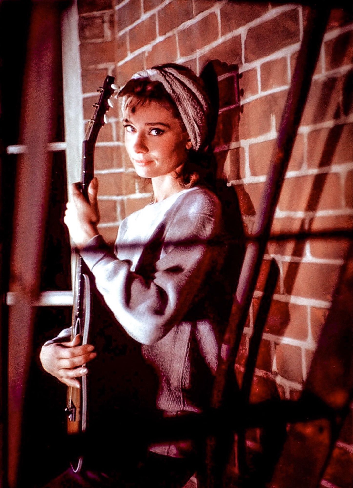 Audrey in a grey long sleeved shirt and cap, leaning against a brick wall in shadow, holding a guitar up