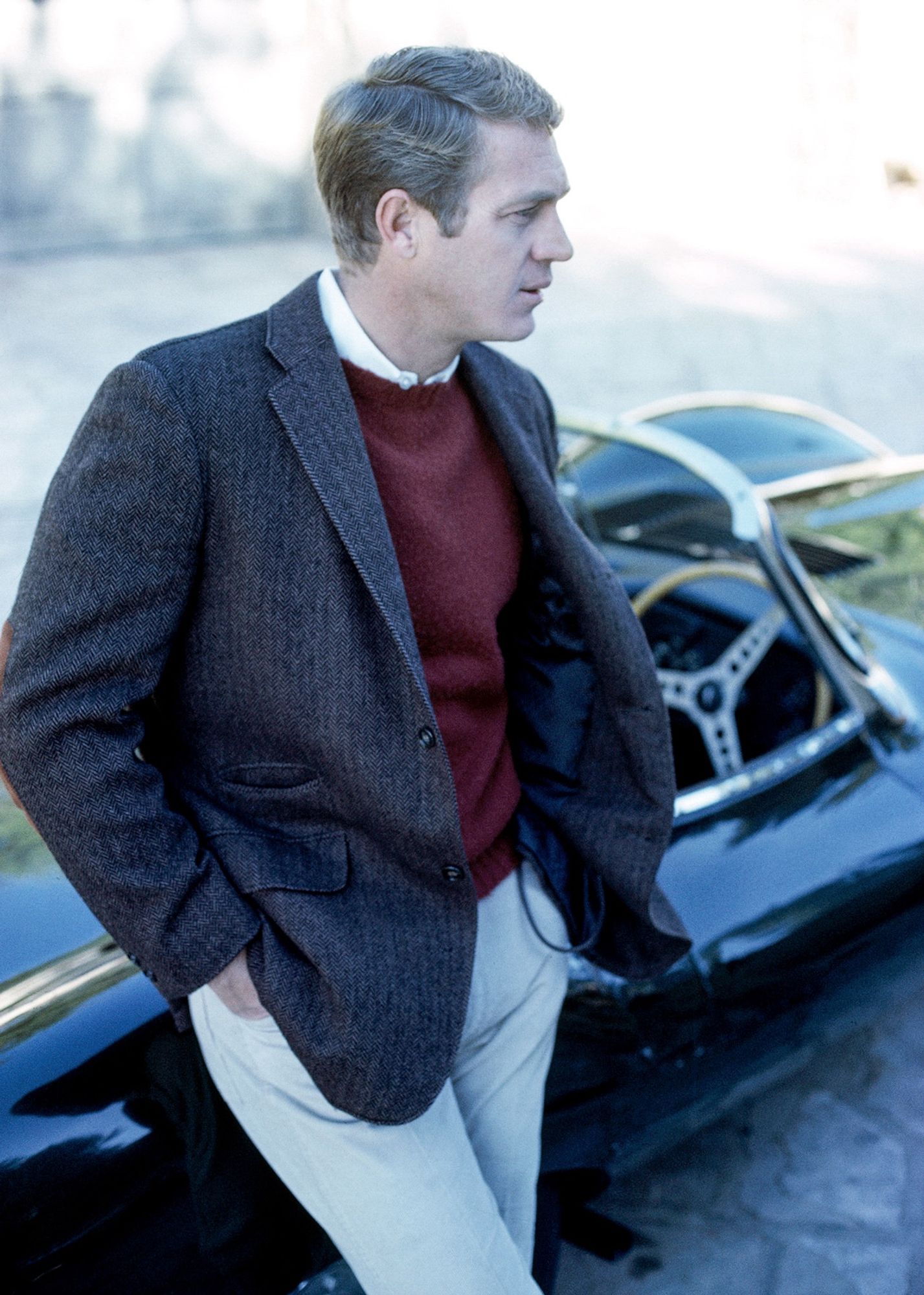 Steve in a burgundy sweater, brown sports coat and white slacks, leaning outdoors against a black roadster, looking thoughtful