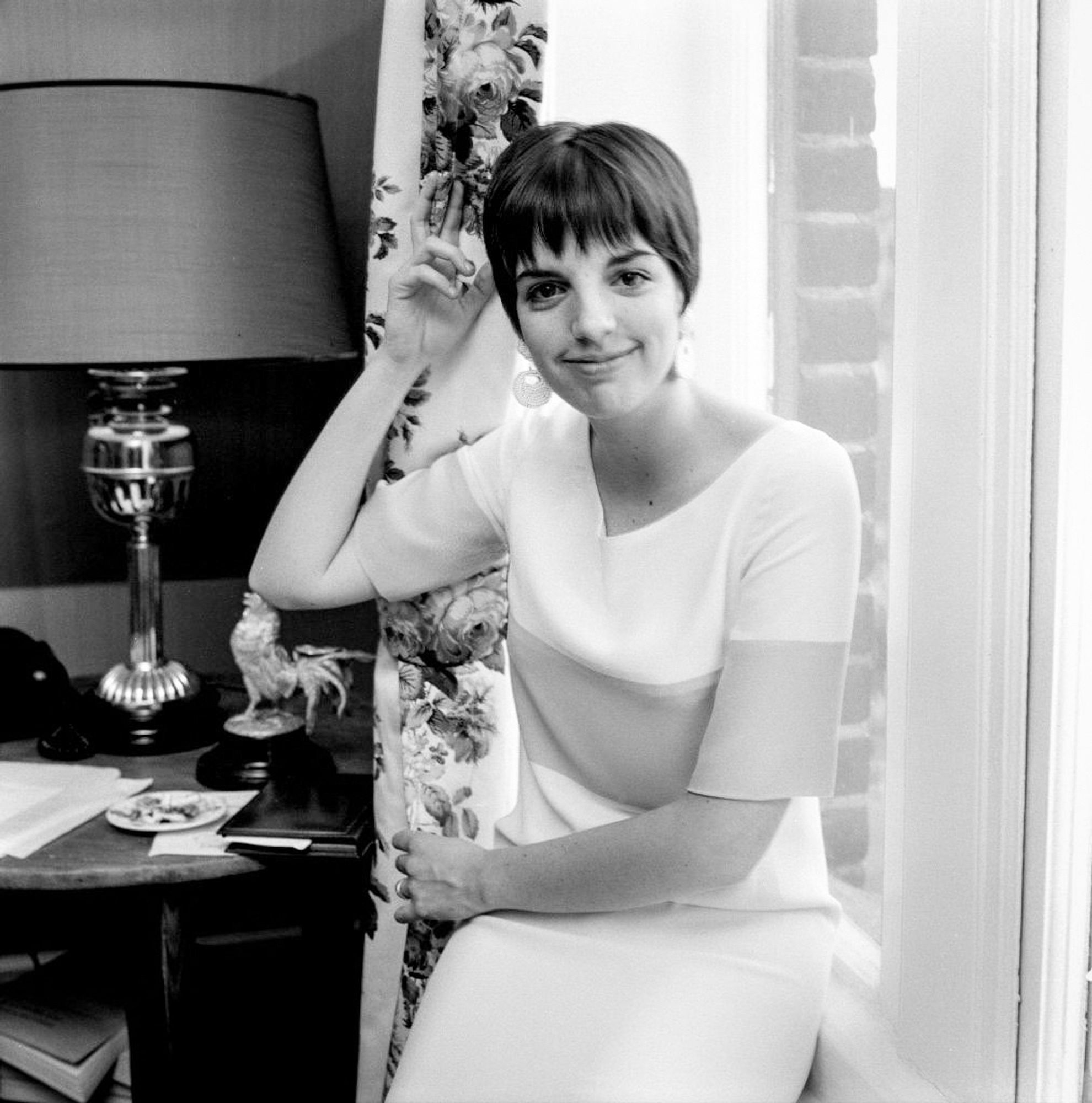 Liza in a black and white photo, sitting in a white dress with a darker colored stripe in the middle. She’s sitting next to a table with a lamp and a rooster statuette on it, and there’s a window to her left. She’s looking straight at the camera and smiling.