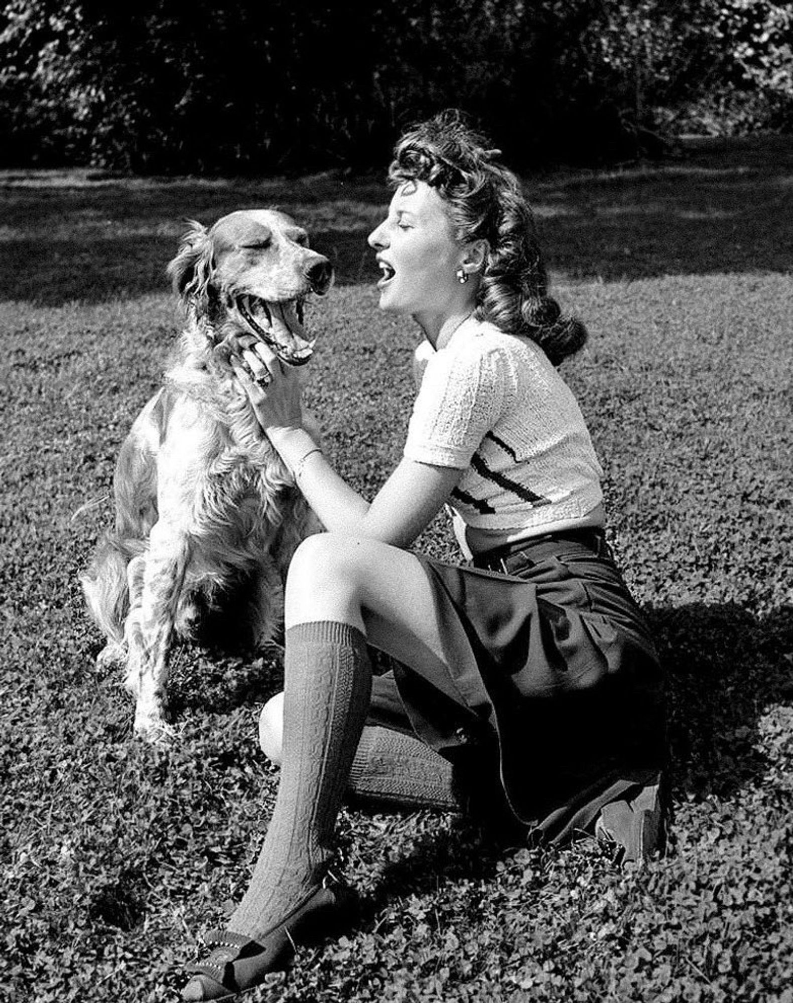 Stany in a short sleeved white knit shirt with vertical stripes, a skirt and long socks, lying on the grass next to a big dog. Both have their mouths opened wide.