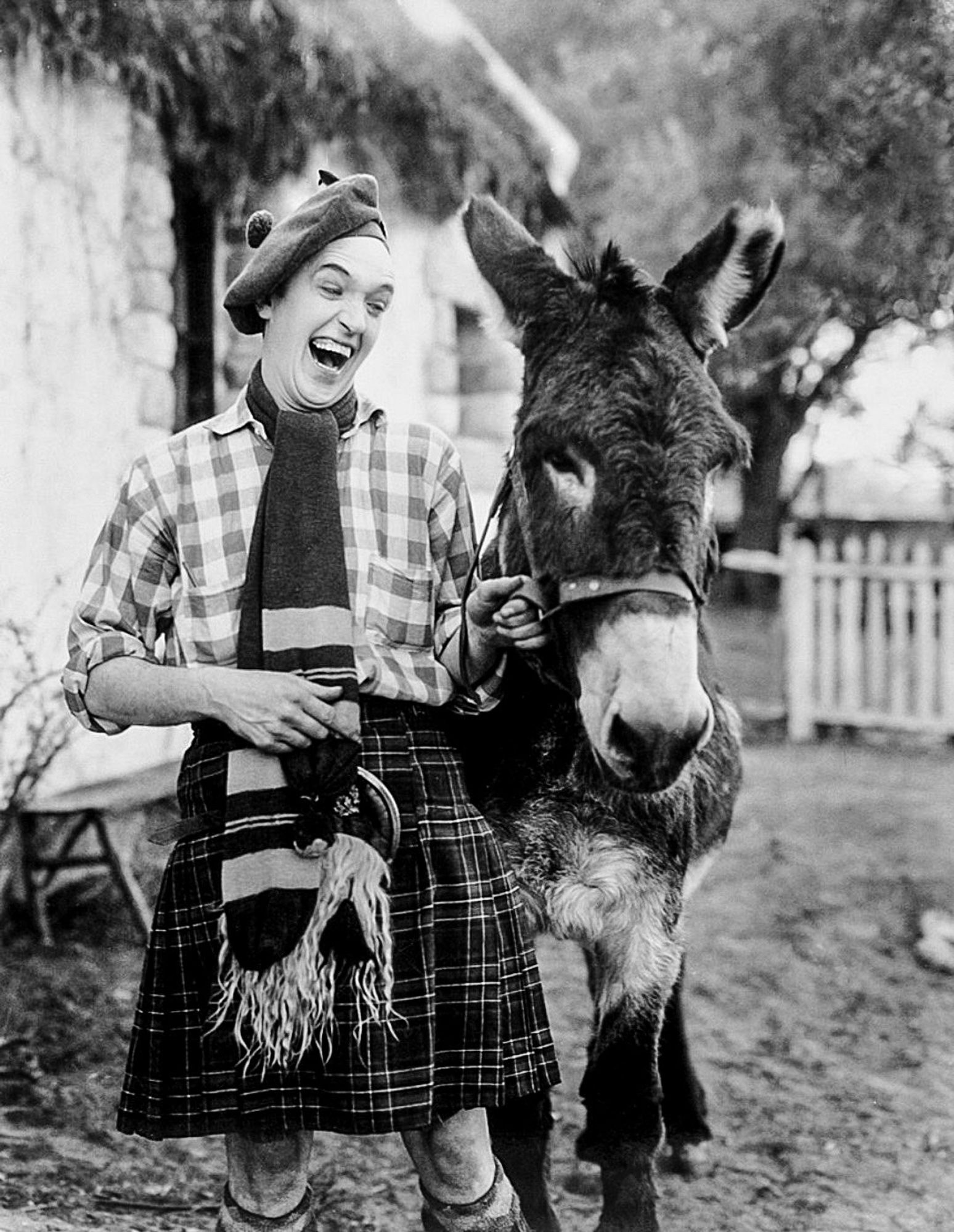 Stan jn a plaid shirt with rolled up sleeves, an oversized striped scarf tied around his neck, and a kilt. He has a beret on his head and is laughing. He’s holding the reins of a donkey, and they’re both standing outside of a house.