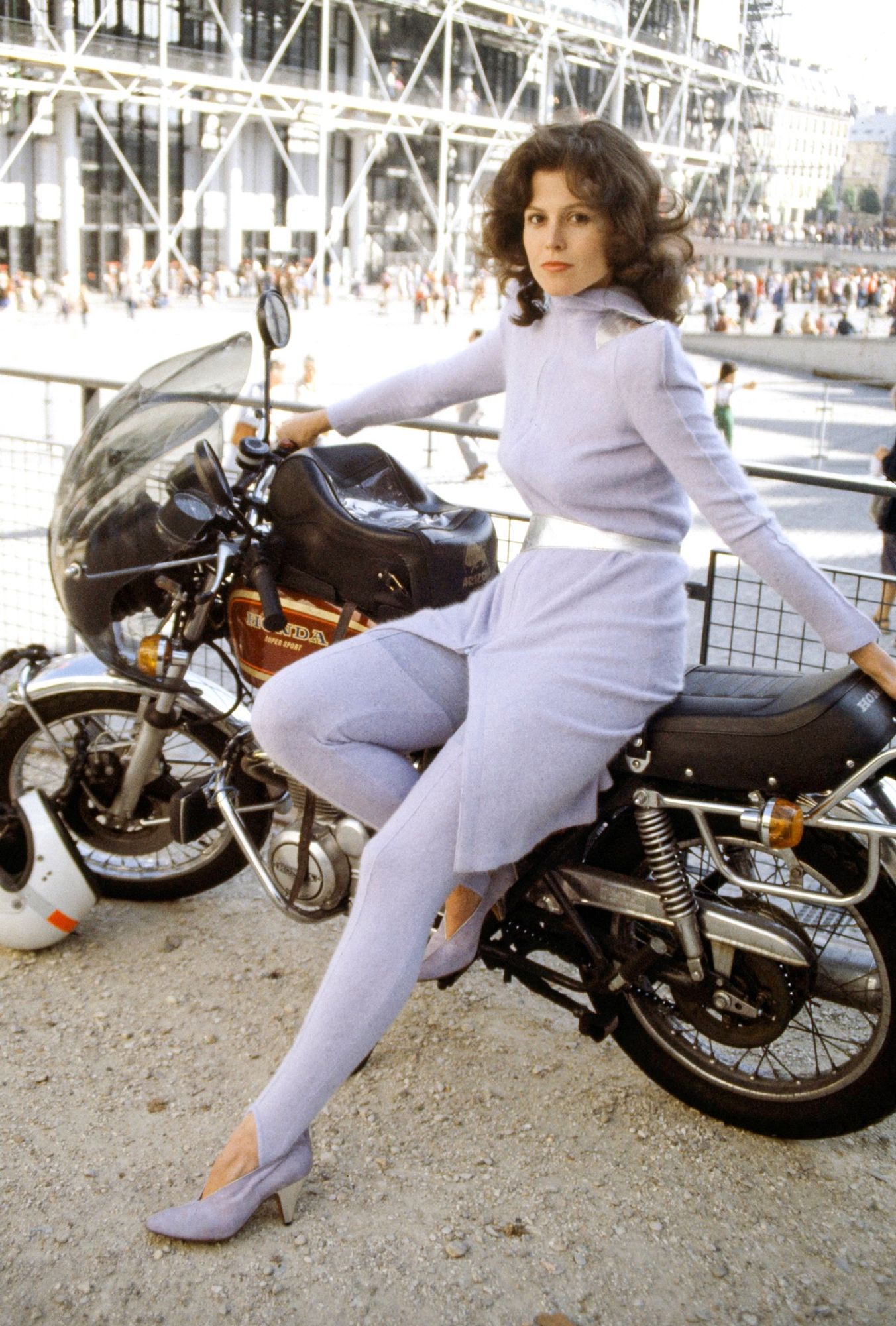 Sigourney in a light purple/violet dress, with matching stockings, and a silver belt. She’s posing in front of a Honda motorcycle in front of the Centre Pompidou