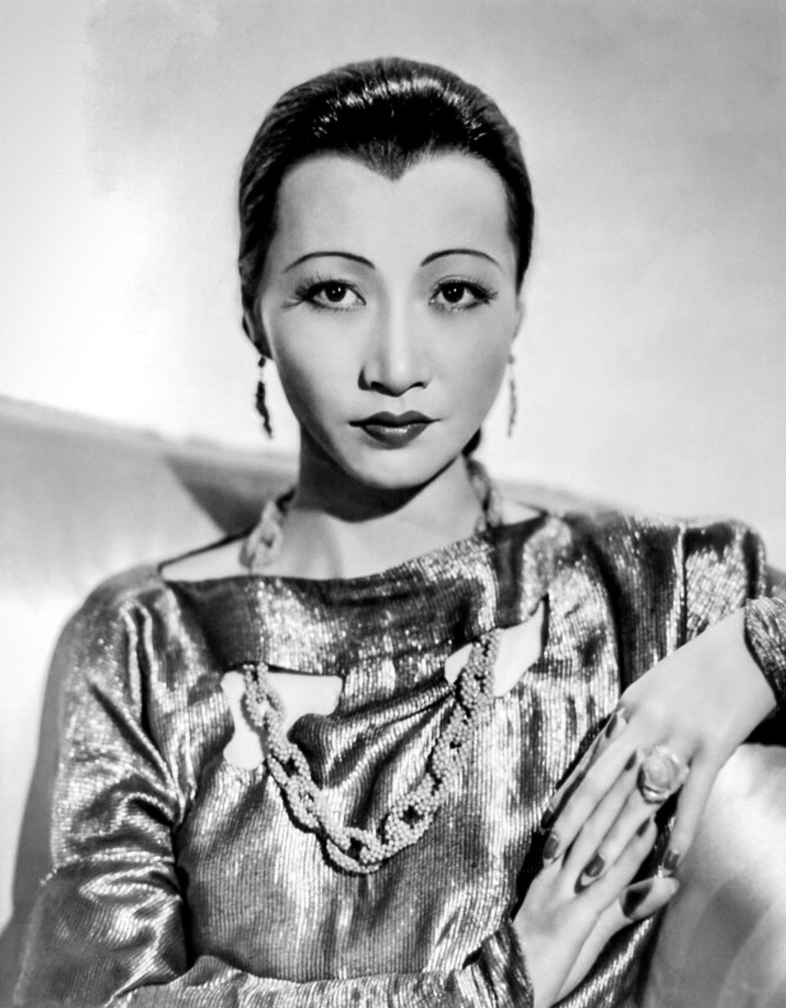 Anna in a sparkling dress, a bejeweled chain link necklace, and leaf shaped earrings, stands with her fingers interlocked.