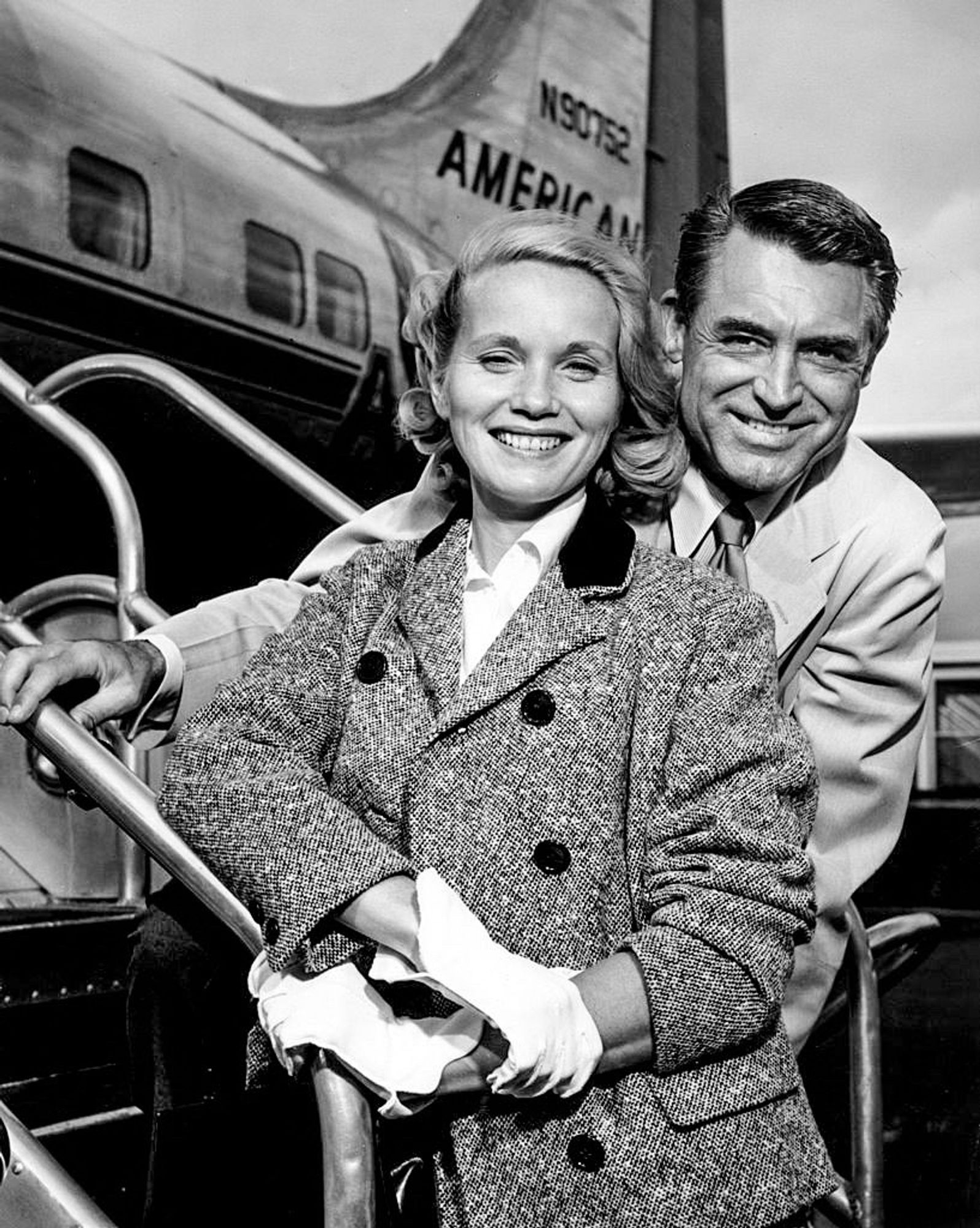 Eva in a white dress shirt and white gloves, with a big knit coat with black buttons. Behind her, and hugging her, is Cary wearing a white suit. Both are smiling broadly, and standing in front of an airplane from American Airlines 