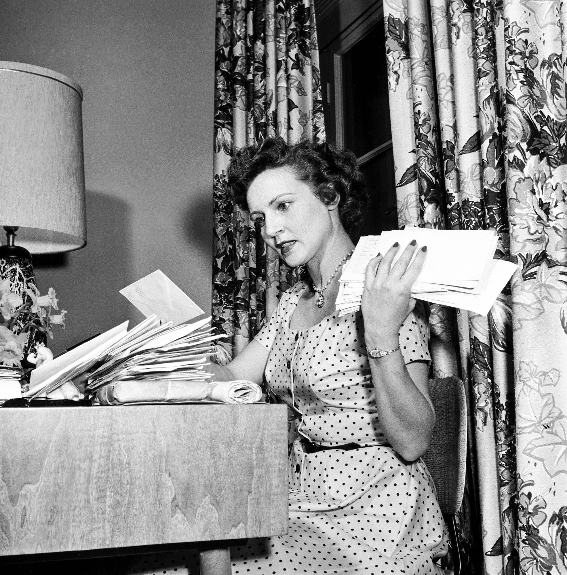 Betty in a white polka dot dress, sitting on a chair at a high wooden table. She has many envelopes in front of her, and she’s holding many more envelopes in her left hand. There’s a lamp on the table and curtains with a flower pattern behind her.