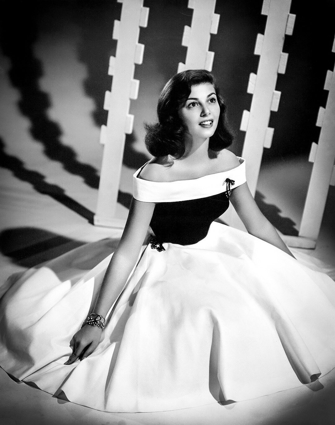 Pier in a black and white dress (with a white band on top, a black torso and a broad white skirt). She’s sitting on the floor in front of an unusual white ridged sculpture, with alternating teeth, and she’s looking up towards the ceiling.