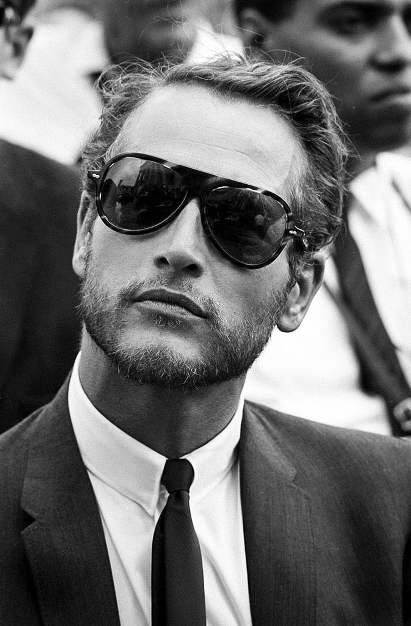 B&W head and shoulders shot of Paul in a suit, standing in a crowd, with sunglasses and a thin beard.