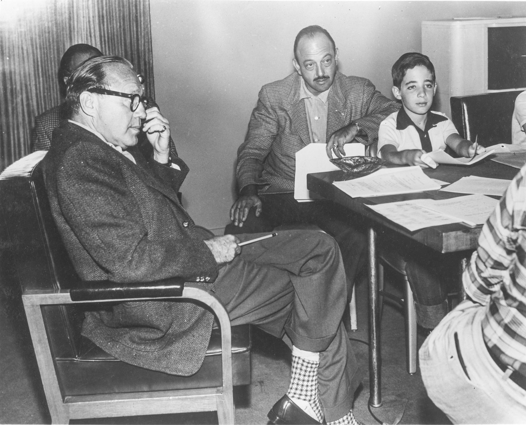 Jack, Mel, and Harry sitting at a table with other people whose faces cannot be seen. Jack and Mel are wearing suits, Harry in a white and black collared shirt. There’s a variety of paperwork on the table.