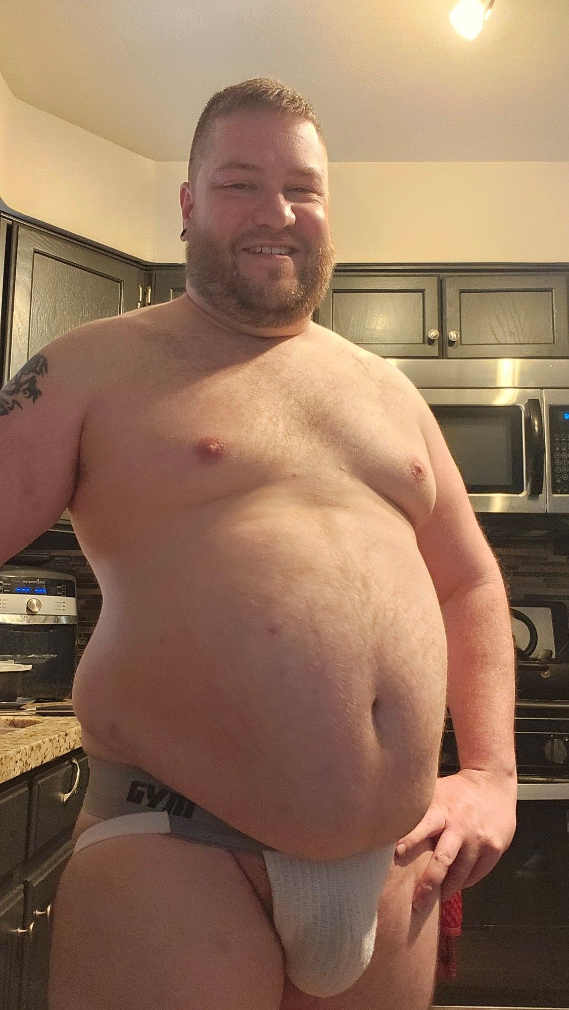 Chubby man standing in a kitchen in a white jockstrap.