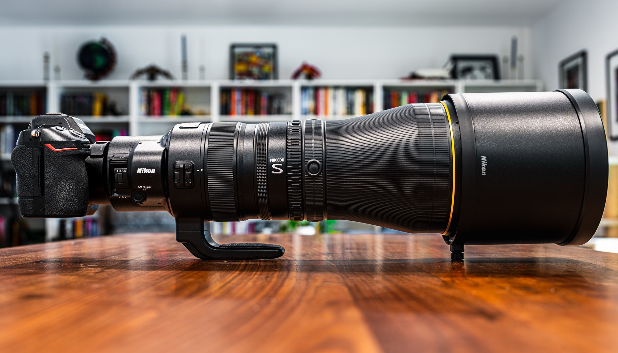 An enormous telephoto lens sits on a wooden tabletop surface. The setup is viewed in profile, with the professional camera body attached to one end looking comically small.