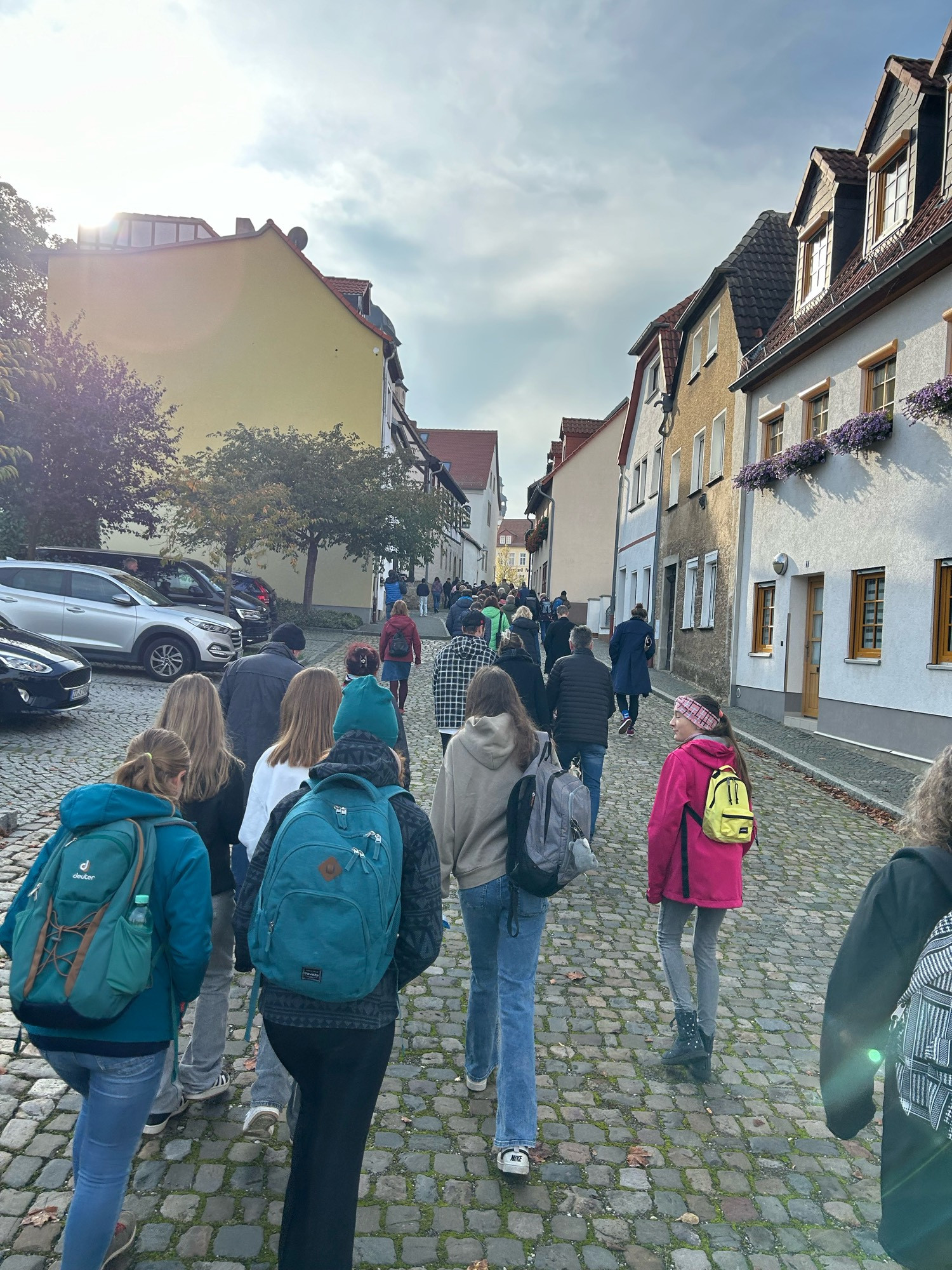 Viele Menschen laufen auf einer Straße einen Anstieg hinauf. Sie tragen bunte Kleidung. Rechts finden sich Stadthäuser, links Autos.