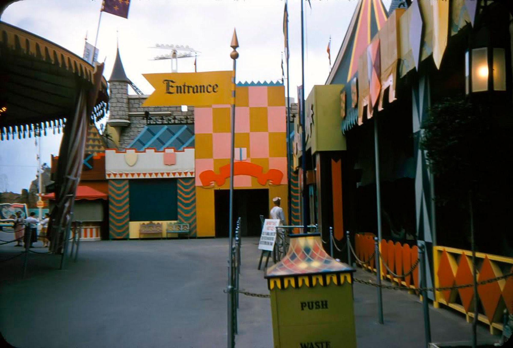 A colorful entrance to an amusement park area, featuring a prominent sign reading "Entrance." The vibrant, patterned buildings are flanked by decorative flags and barriers. A man is seen walking toward the entrance, and there's a trash can labeled "PUSH."