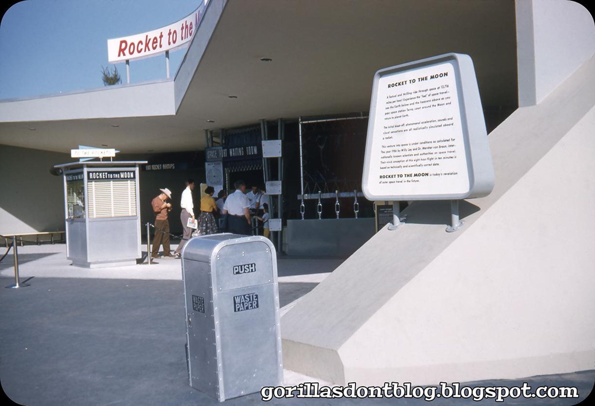  A amusement park ride entrance featuring a sign that reads "Rocket to the Moon." In front, there are ticket booths and a wastepaper can. A small crowd of visitors is gathered, some reading brochures. The architecture has a mid-century modern design.