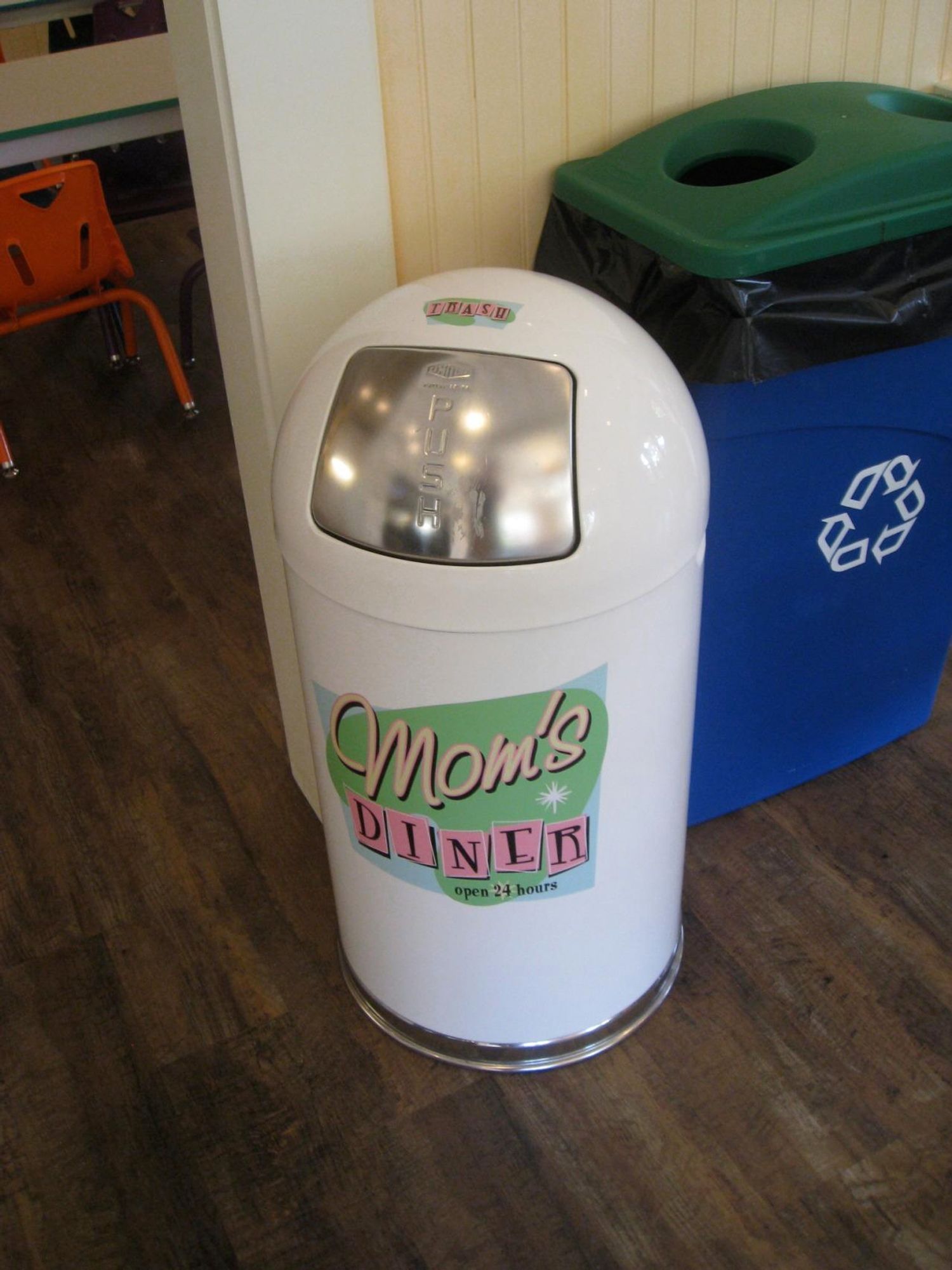 A white trash can labeled "Mom's Diner" with a circular silver push-top lid, positioned next to a blue recycling bin. The setting has wooden flooring and hints of a diner ambiance. 