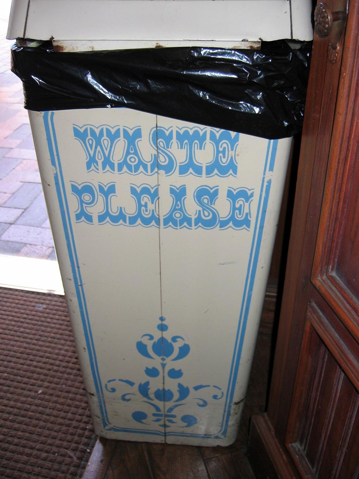  A vintage trash can with a decorative design, labeled "WASTE PLEASE" in blue text, and covered with a black trash bag at the top. The bin is positioned beside a wooden structure, likely within an indoor setting. 