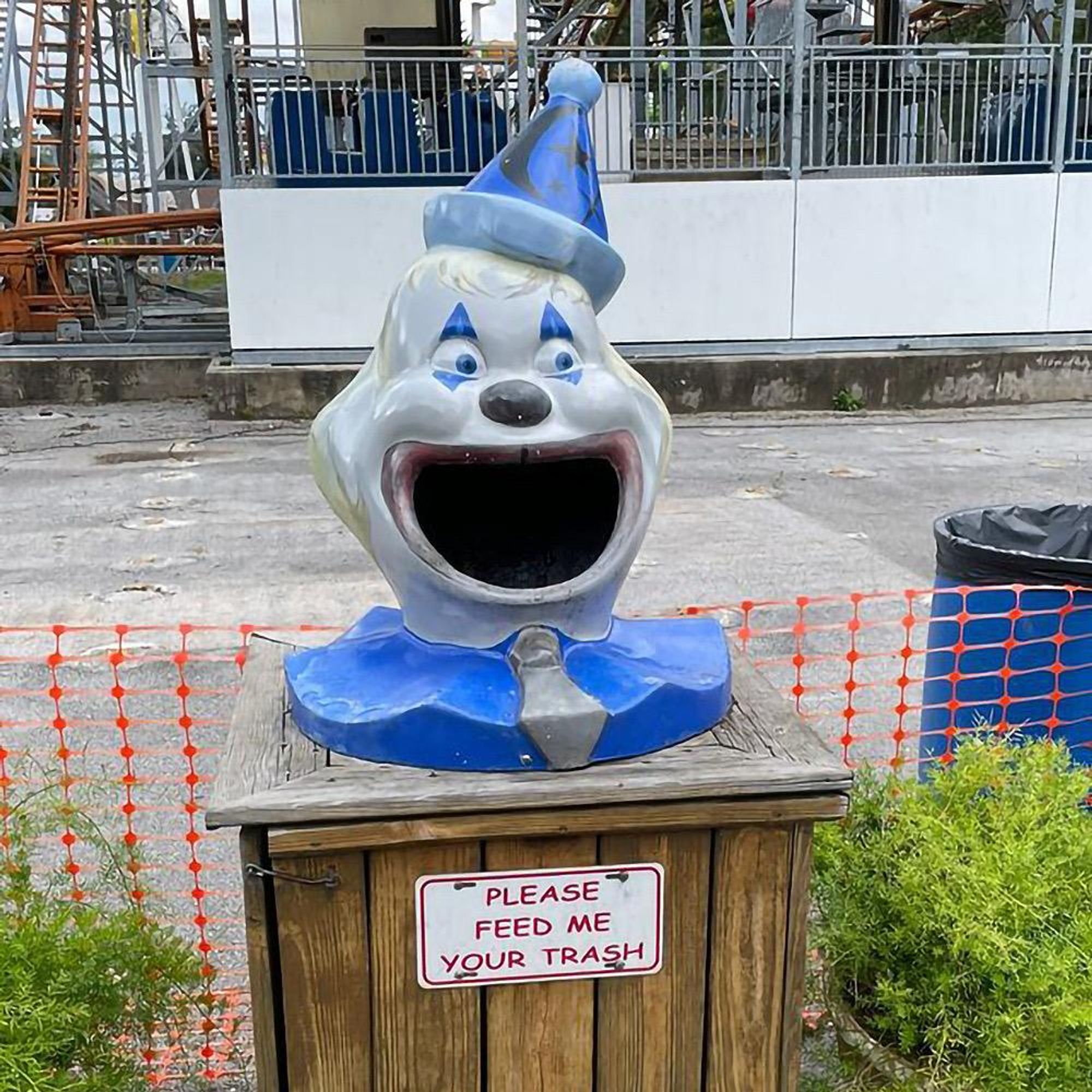 A clown-shaped trash can with a wide-open mouth, wearing a blue party hat and bow tie. The wooden base features a sign that reads, "PLEASE FEED ME YOUR TRASH." 