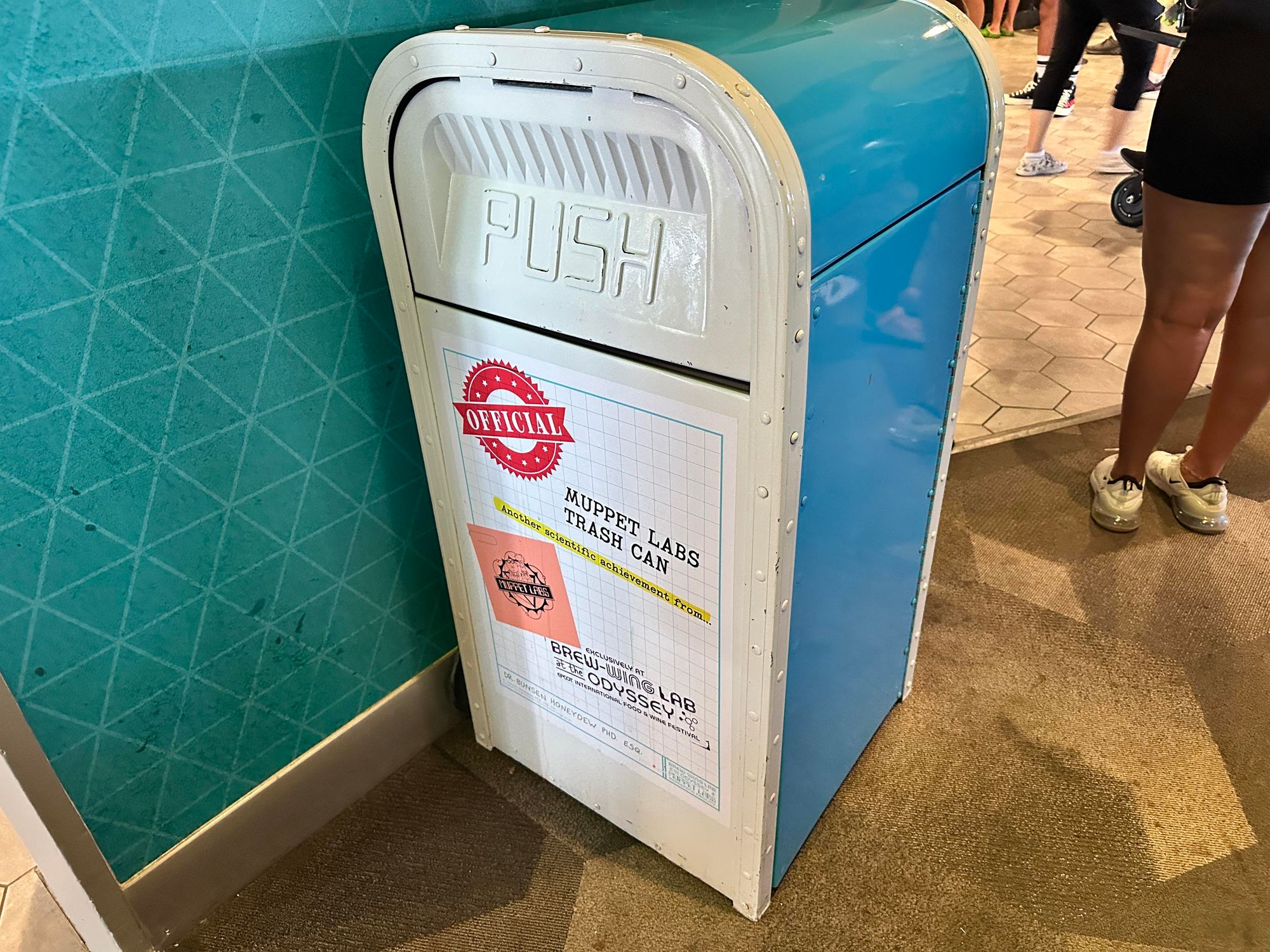 A blue and white trash can labeled "Muppet Labs Trash Can" with an "Official" stamp. It's placed against a textured teal wall in a busy indoor area. 