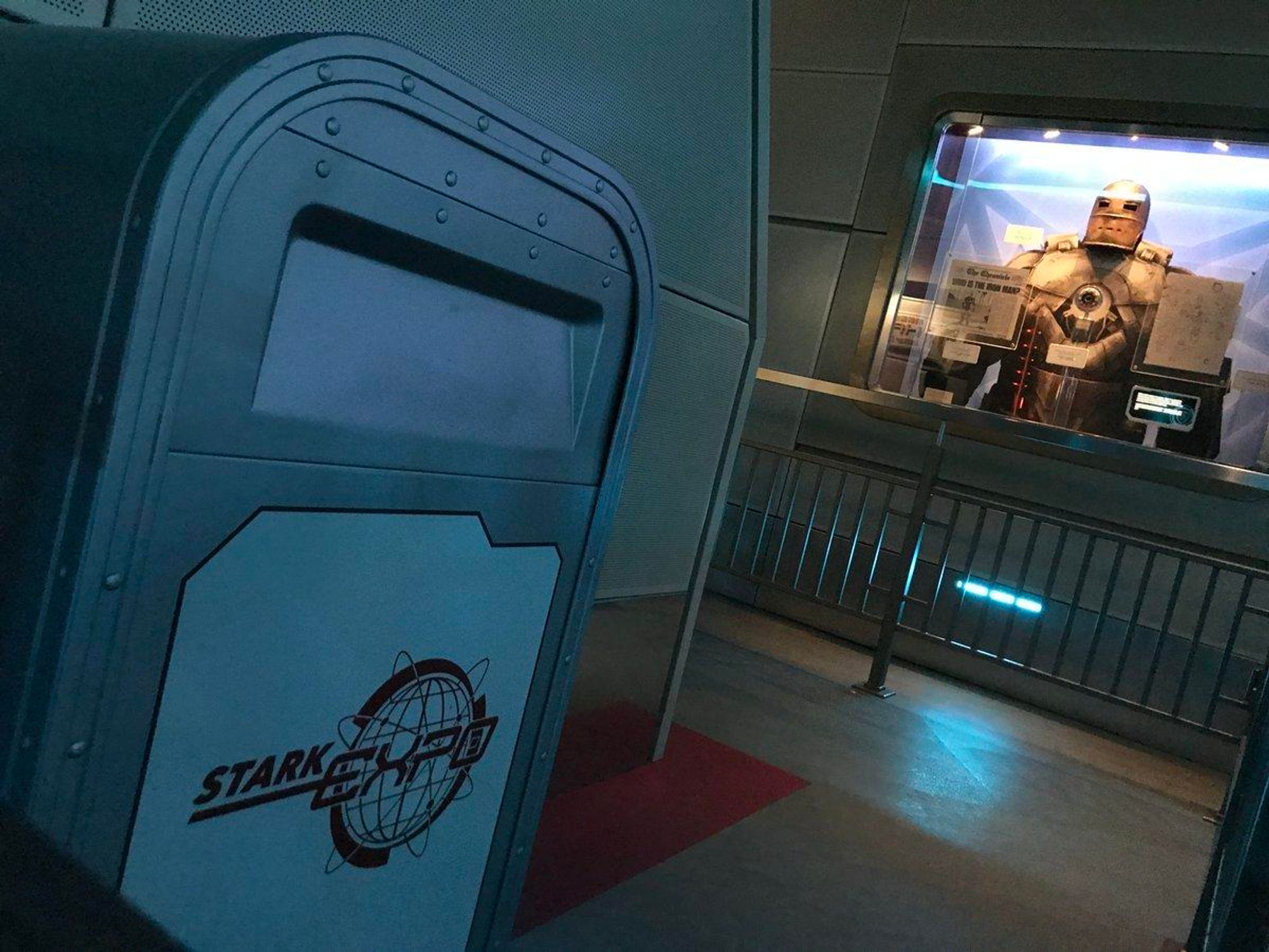 An indoor display featuring a Stark Industries trash can in the foreground and a glass case showcasing an Iron Man suit in the background. The setting has a sleek, futuristic design. 