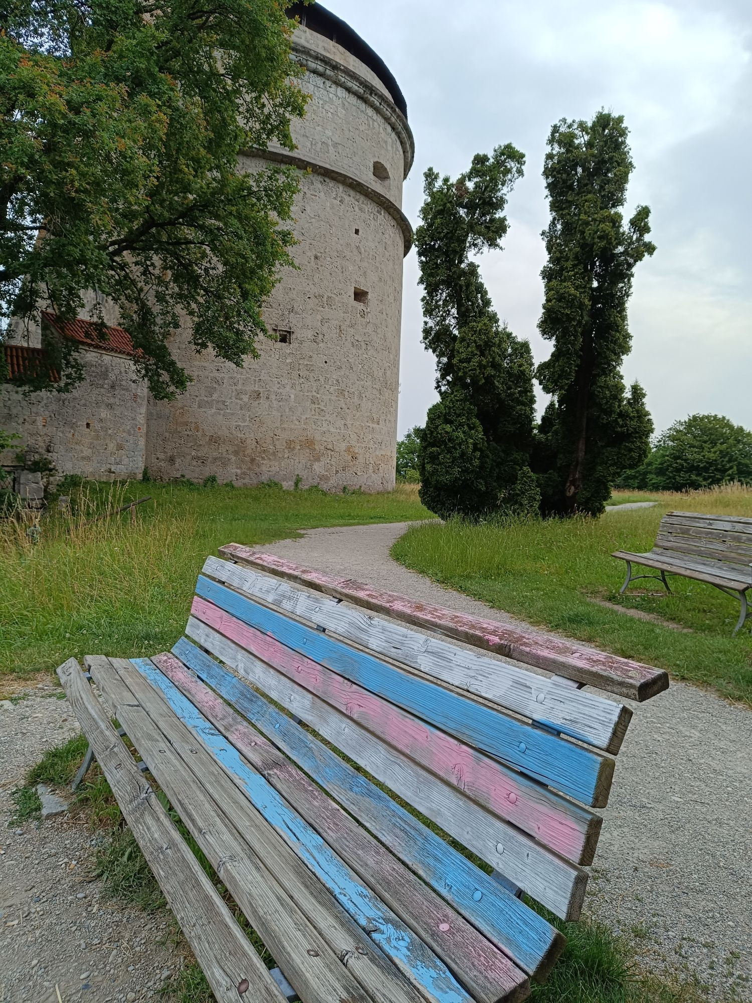 Holzbank in den Farben der Trans-Flagge rosa hellblau weiß. Im Hintergrund mittelalterliche Wehranlage