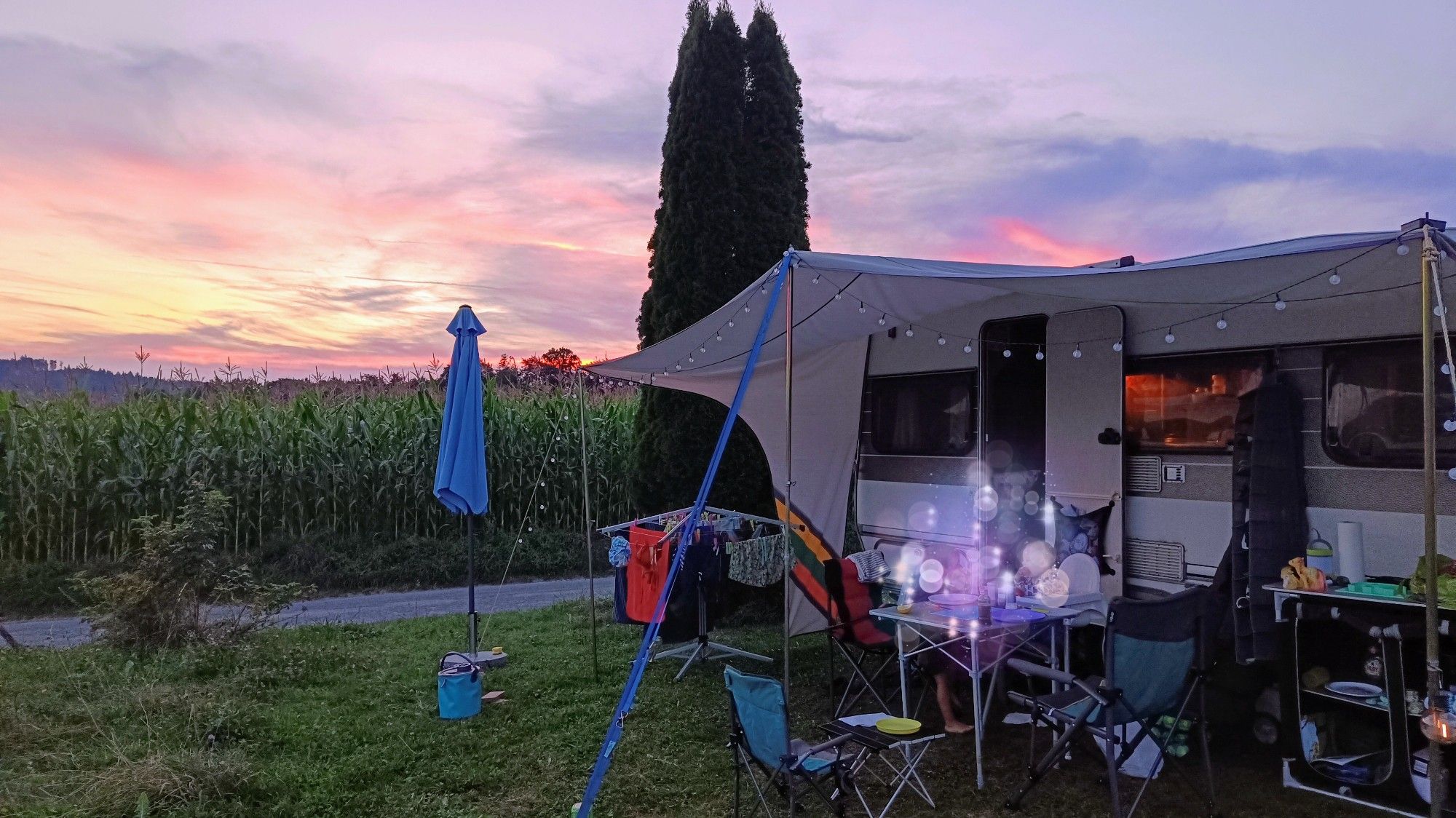 Wohnwagen mit Vorzelt und Kram steht am Rande eines Maisfeldes, im Hintergrund ein schöner Sonnenuntergang