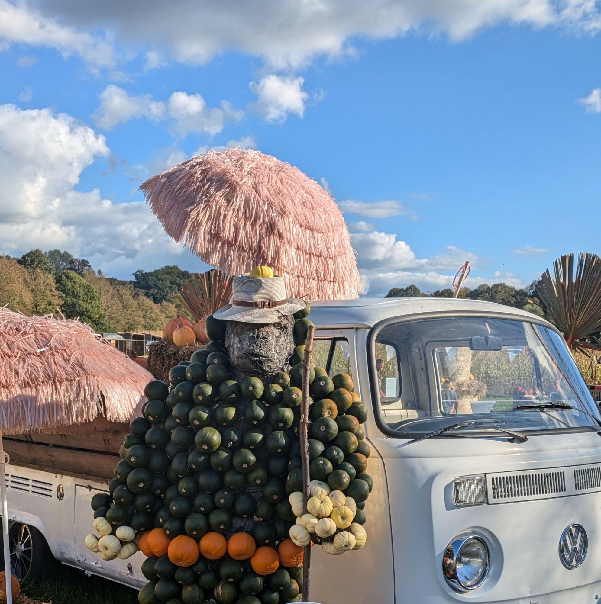 Figur mit Hut, aus kleinen grünen und orangenen Kürbissen zusammengesetzt, lehnt lässig an einem VW Bulli 