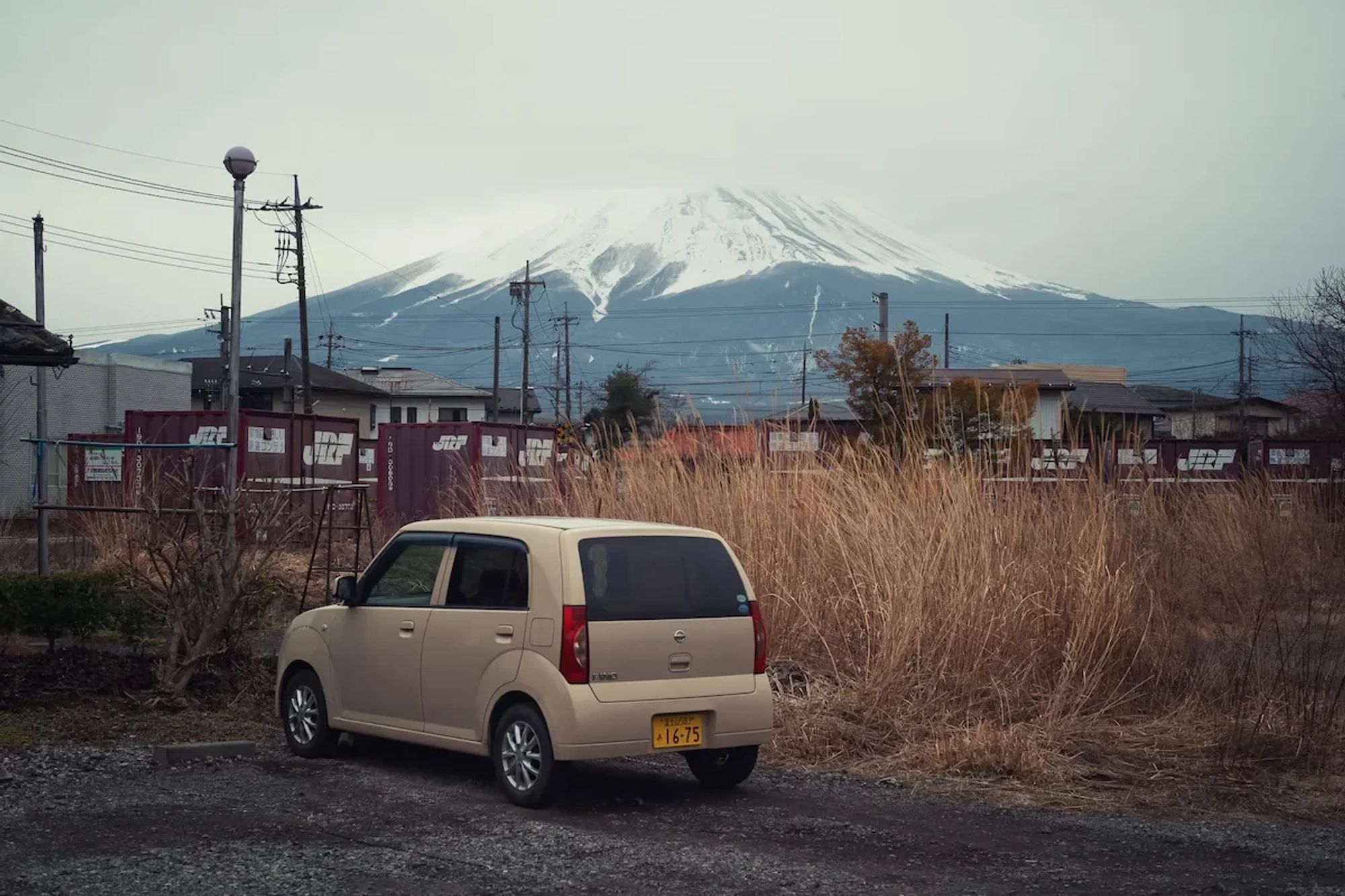 A car is parked with houses in the background. Beyond the houses Mount Fuji is seen with plenty of snow on it.