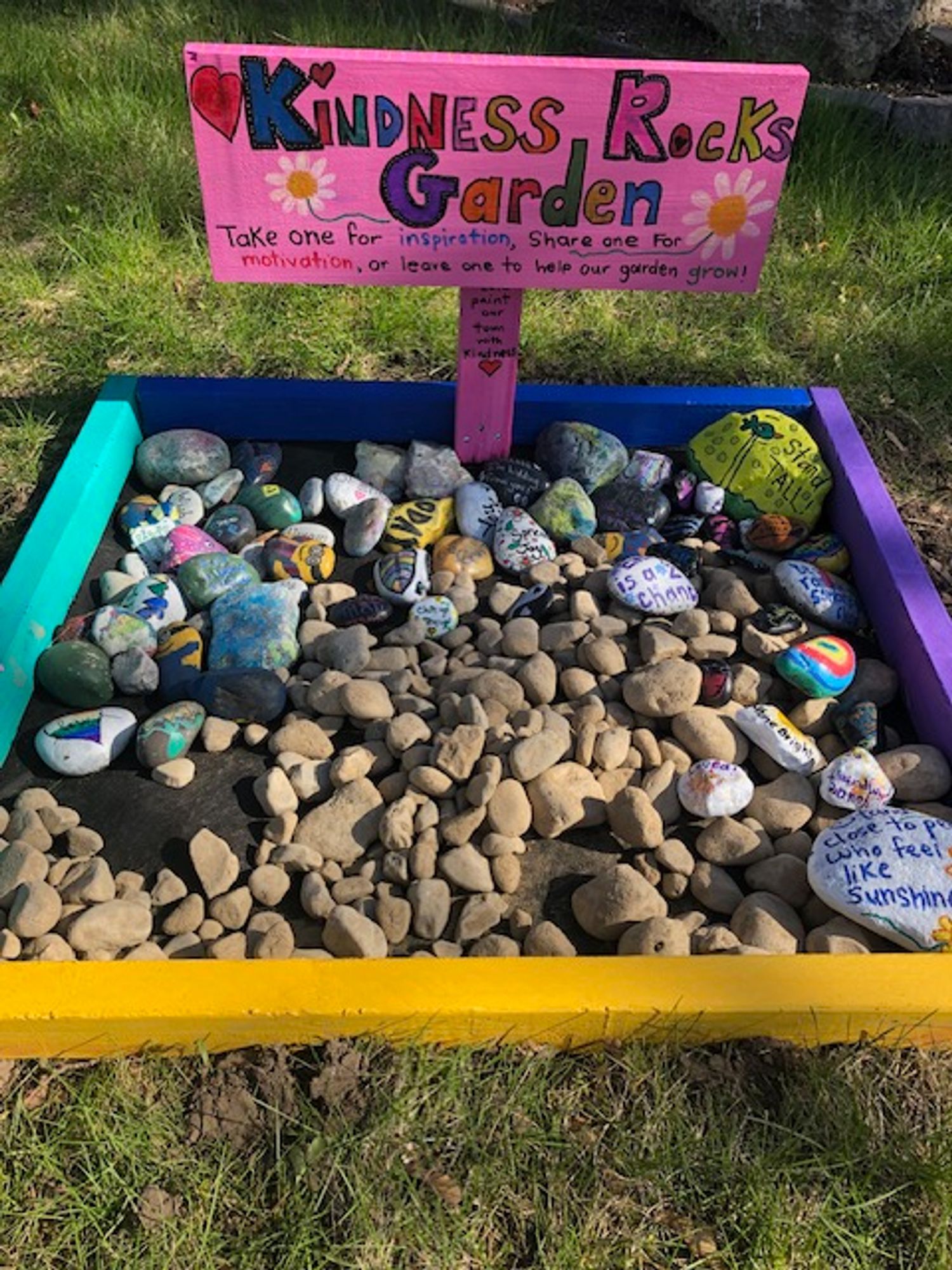 The letters, Kindness Rocks Garden, are in different colours on a  wooden sign painted pink with a couple of daisies on it and a red heart. Underneath the name of the garden are the following instructions: "Take one for inspiration, share one for motivation, or leave one to help our garden grow!" The sign, attached to a flat wooden post also painted pink, has the following inscription: "We paint our town with kindness." A red heart is drawn underneath the inscription. In the colourful wooden 'garden' box, painted purple, yellow, and two shades of blude are numerous decorated stones as well as stones awaiting decoration and/or inscription. One of the painted stones in the bottom right has the following inscription: "Stand close to people who feel like sunshine."