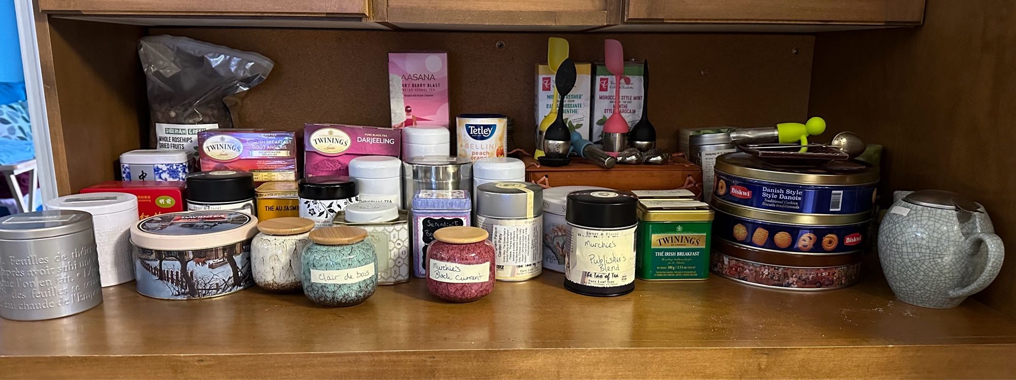 Shelf full of teas, most in canisters of various sizes, including repurposed chocolate and cookie tins.