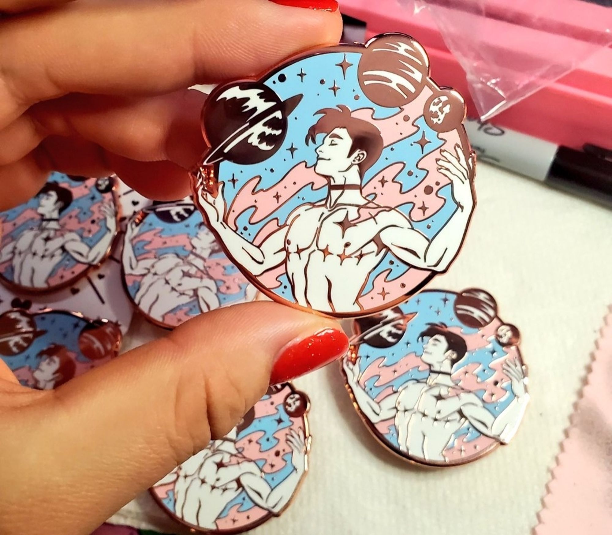 Photo of a hard enamel pin that depicts a triumphant trans masculine figure surrounded by planets and pink and blue space dust. His top surgery scars are proudly displayed.