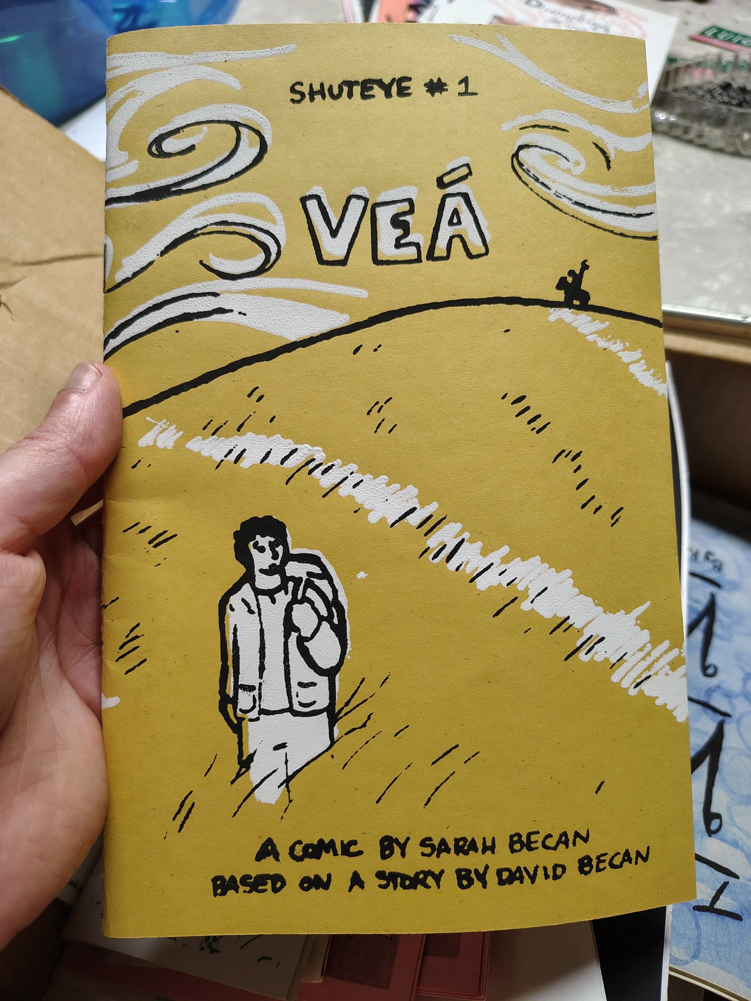 Elliott's hand is holding a comic zine by Sarah. The cover is goldenrod yellow with a man standing in a field as the wind whips around him. The illustrations are screenprinted in black and white. The title is Shuteye #1: Vea