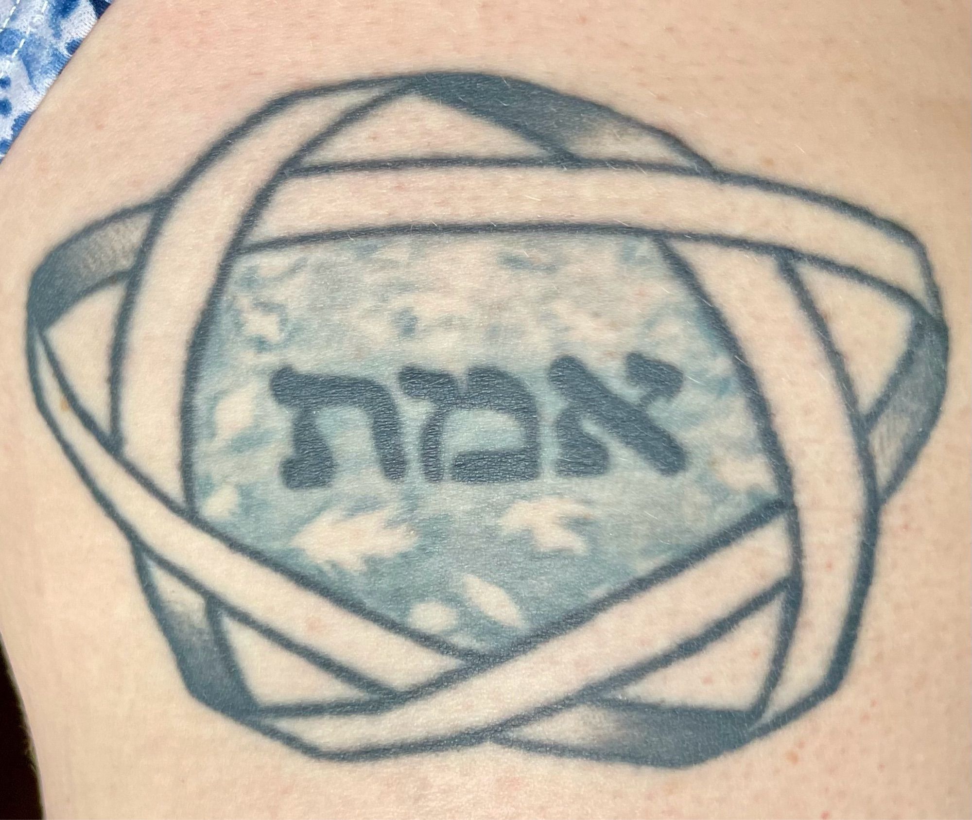 A monochrome tattoo of a five-pointed Mobius strip surrounding the Hebrew word “emet,” with leaves floating in a water surface in the background.