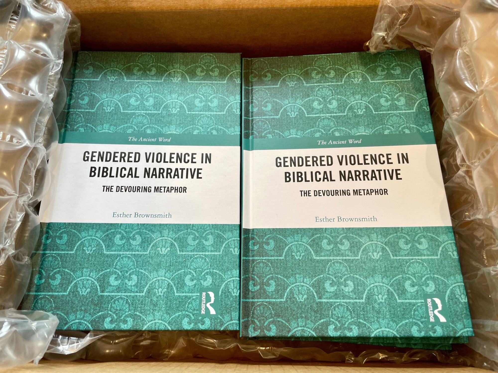 A cardboard box with bubble wrap surrounding books with a green patterned cover and the title "Gendered Violence in Biblical Narrative: The Devouring Metaphor," by Esther Brownsmith.