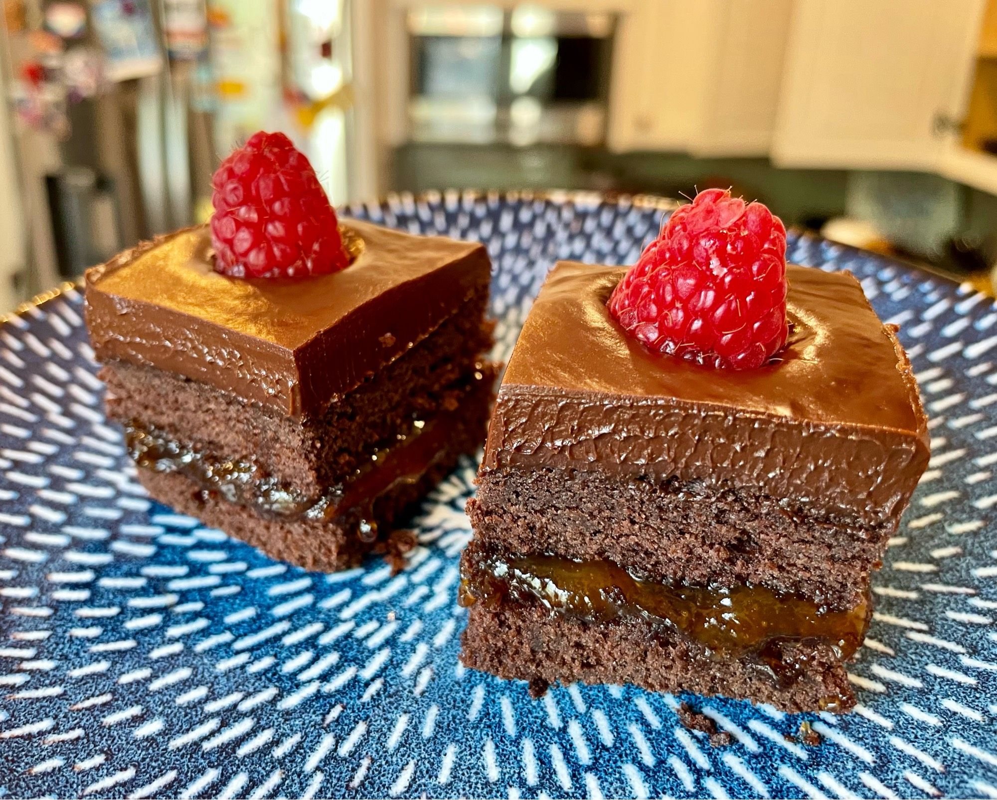 A photo of two cake squares on a blue plate. The cakes are layered chocolate, topped with a fresh raspberry.