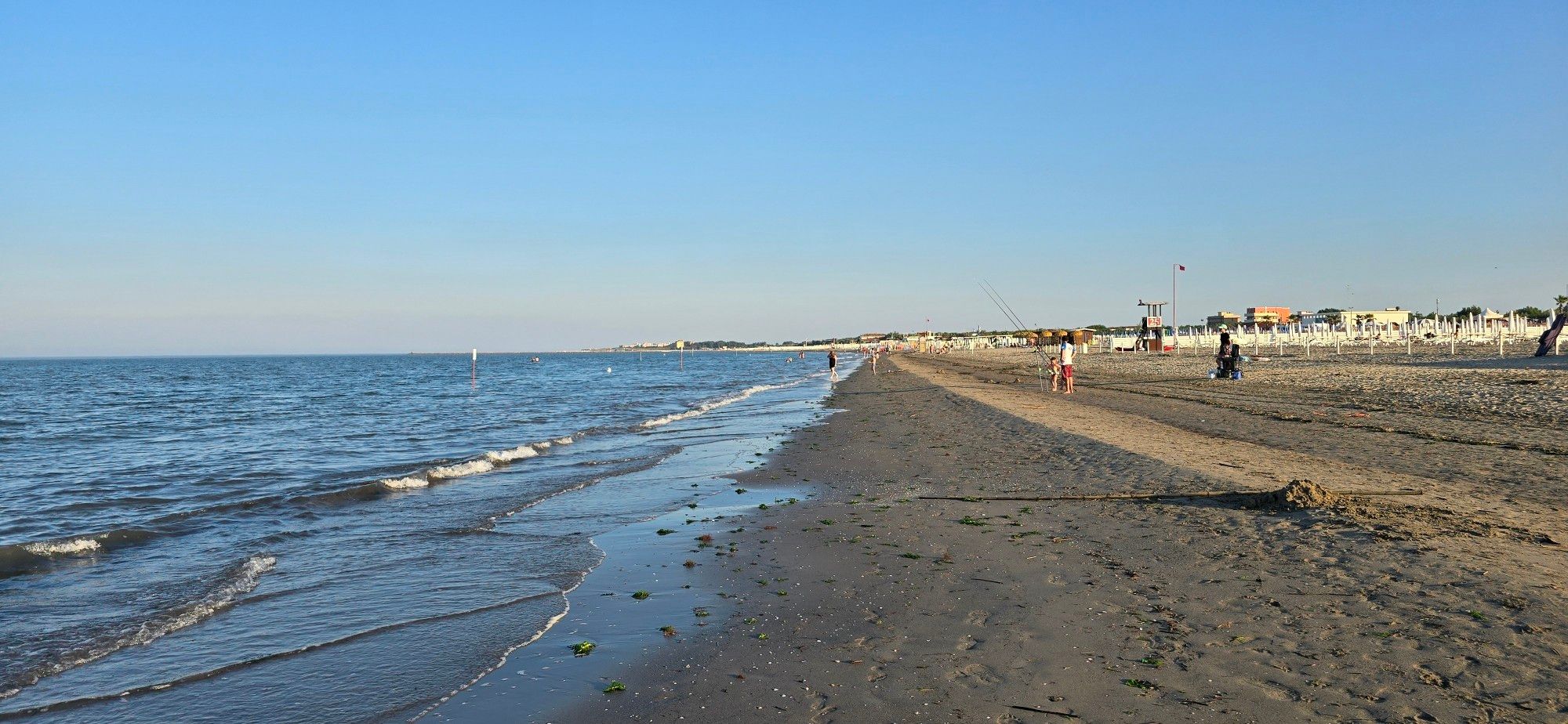 Blick über den Strand 
Links das Meer und kleine Wellen. Rechts Sandstrand und einzelne Menschen. In der Ferne rechts ein paar Gebäude und ein Rettungsschwimmerturm.