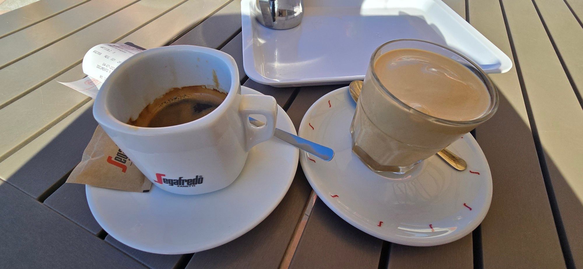 Links im Bild eine Tasse Espresso mit einer Untertasse mit einem Löffel und einem Päckchen Zucker
Rechts im Bild ein Glas mit hellbraunem Eis auf einer Untertasse
Im Hintergrund ein Tablett
Eine Tischplatte aus einzelnen Brettern als Untergrund