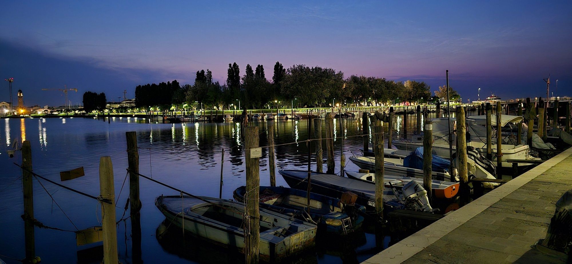 Blick im Dunkeln auf ankernde Boote im Vordergrund und Baumsiluetten im Hintergrund und sich spiegelnde Lichter