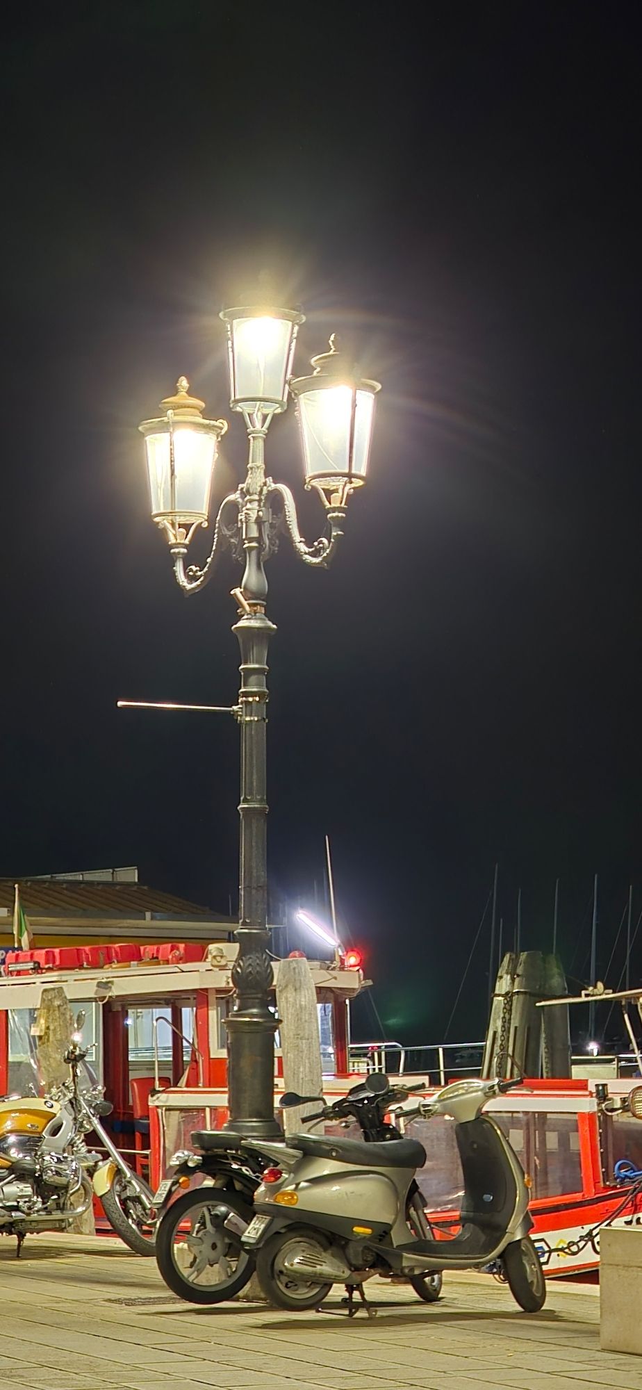 Eine 3 armige eingeschaltete Straßenlaterne im Dunkeln, darunter geparkte Motorroller und ein kleines rotes Schiff