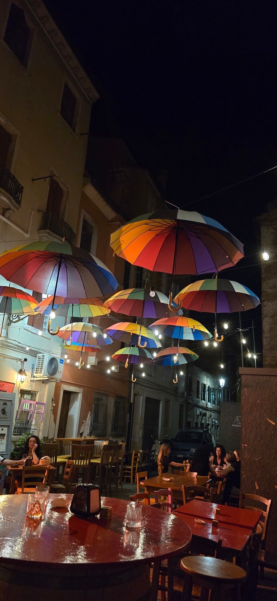 Foto im Dunkeln: Tische, Stühle und Menschen im Restaurant, darüber regenbogenfarbige Schirme aufgespannt mit Lichterkette dazwischen