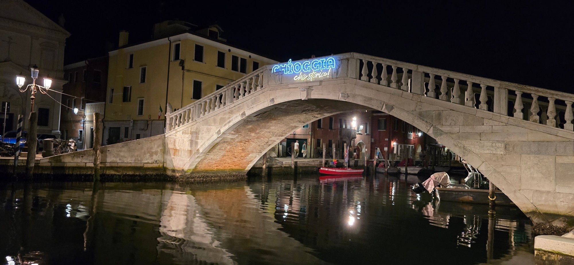 Die Vigo-Brücke mit der Aufschrift "Chioggia che storia!" 
Eine weiße Brücke im Dunkeln
Links davon eine Straßenlaterne, im Hintergrund eine Fassade
Unter der Brücke spiegelt das dunkle Wasser die Lichter