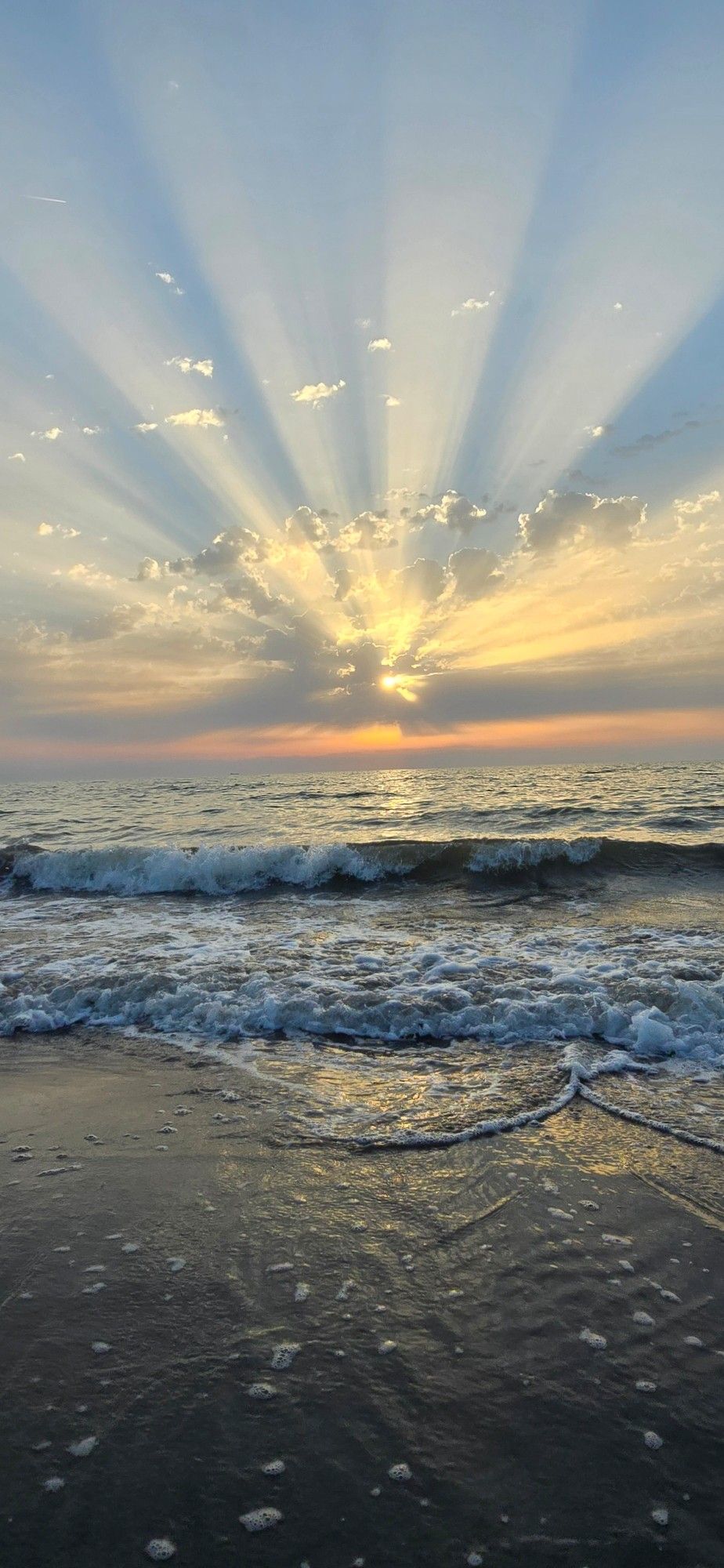 Im Vordergrund das Meer und leichte Wellen
Am Horizont scheint die Sonne durch die Wolken und bildet einen Strahlenkranz.