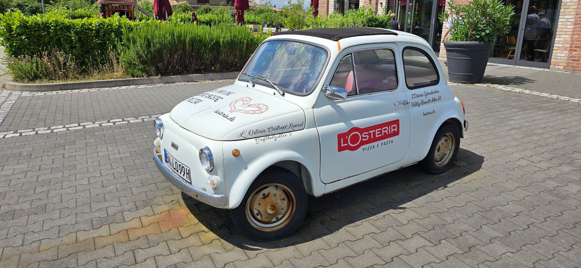 Ein alter weißer Fiat mit Werbung L'Osteria auf einem gepflasterten Parkplatz 
Im Hintergrund Hecken