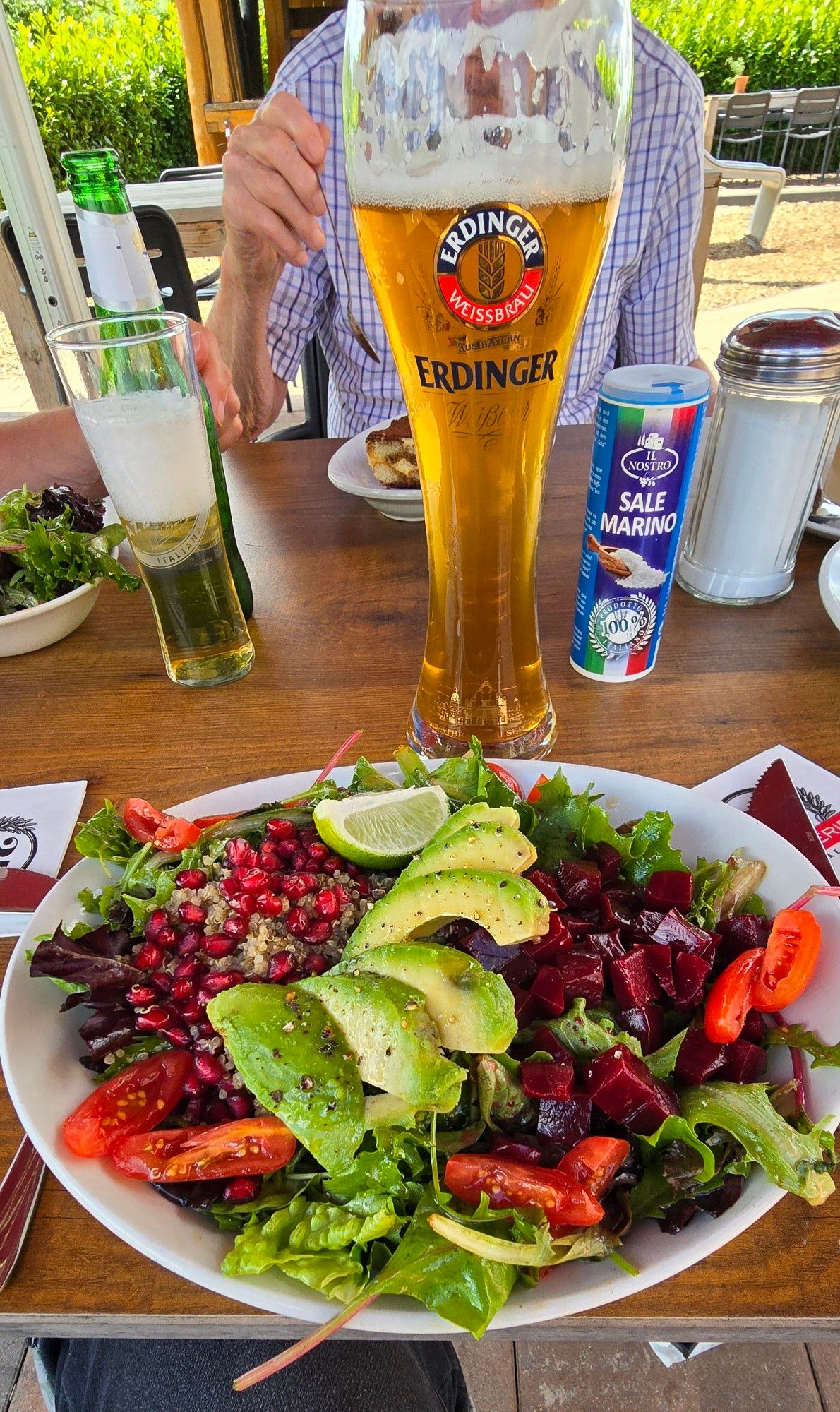 Ein bunter Salatteller u.a. mit Granatapfel und Avocado, ein Erdinger-Glas, italienisches Meersalz, ein Zuckerglas und ein kleines Bierglas stehen auf einer Holztischplatte. Im Hintergrund ist eine Person im karierten Hemd mit einem Löffel in der rechten Hand erkennbar.