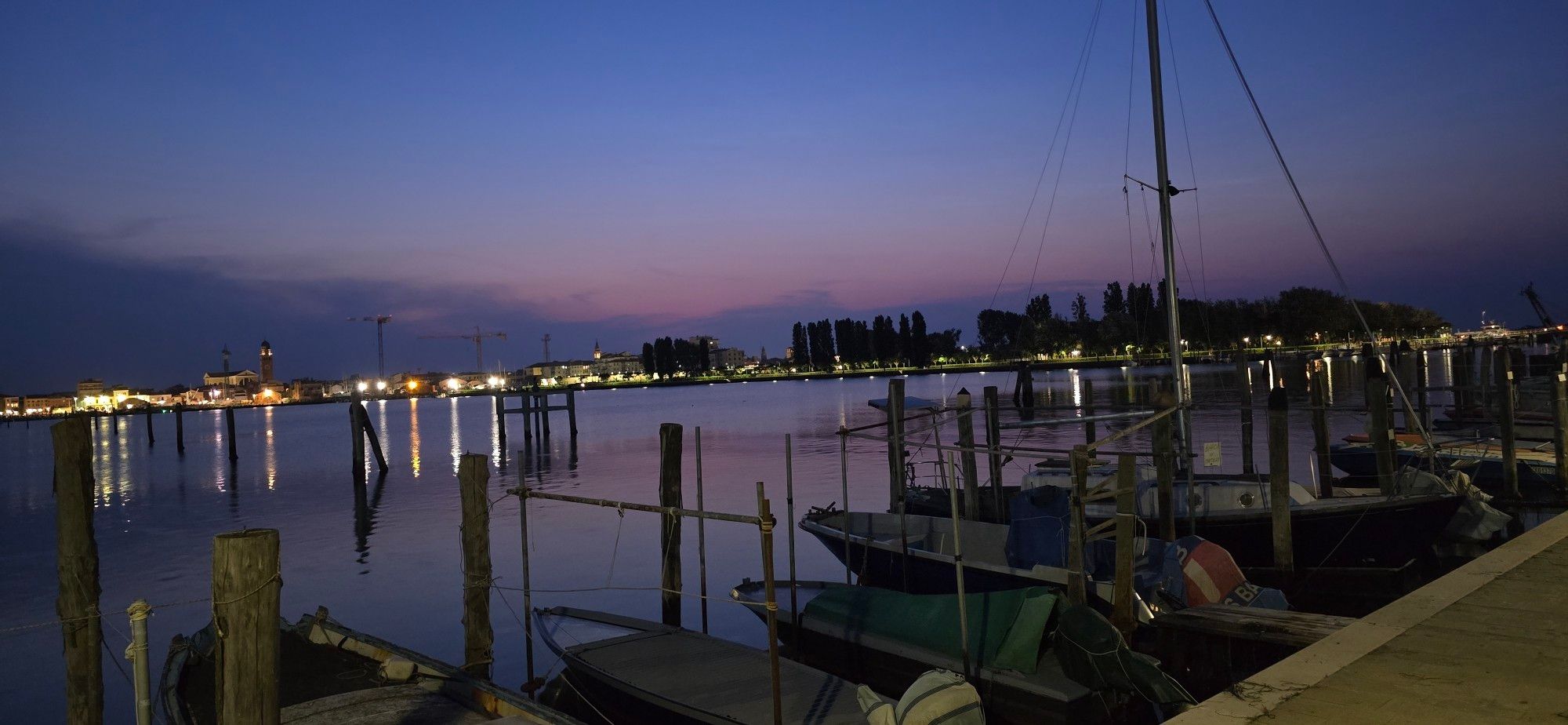 Blick im Dunkeln auf ankernde Boote im Vordergrund 
Im Hintergrund leicht lila Wolken und die Wasseroberfläche spiegelt einige Lichter vom gegenüberliegenden Ufer.