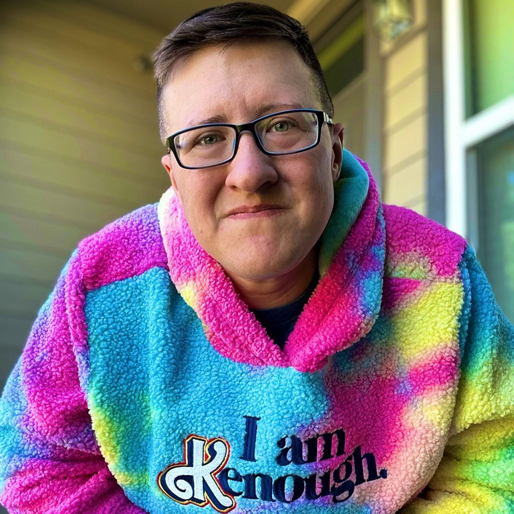 A white androgynous person with black rimmed glasses smiling at the camera.  They are wearing a pink, blue, and yellow tie dye style fleece sweatshirt.  The sweatshirt has the words, “I am Kenough.”