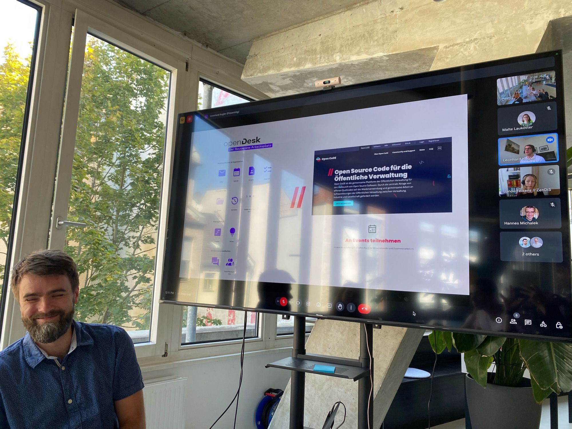 A man with a beard and short hair is sitting and smiling next to a large screen displaying a virtual meeting. The screen shows a presentation slide titled "OpenDesk" and a webpage related to open-source code for public administration. Several participants are visible on the right side of the screen, connected via a video call. The room has large windows with a view of green trees outside, and the setting appears to be a casual office environment.