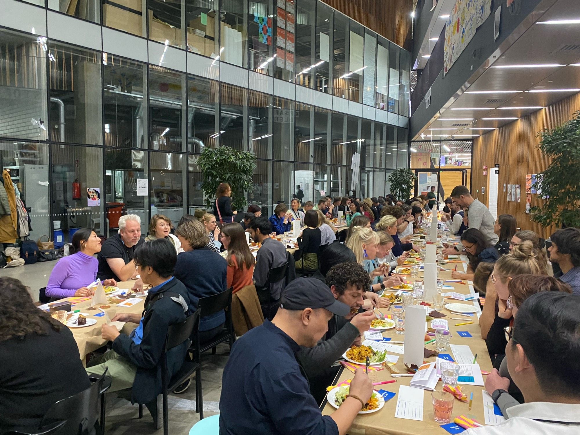 A large group of people sitting at long, communal tables in a modern indoor setting. The space has high ceilings, glass walls, and a well-lit, industrial aesthetic. The tables are arranged in rows, and the participants appear to be engaged in conversation while eating meals. The event seems casual yet organized, with some individuals focused on their food and others talking to their tablemates. There are plants placed around the space, adding a natural touch to the environment. Overall, it appears to be a community gathering or a shared dining experience in a spacious, contemporary venue.