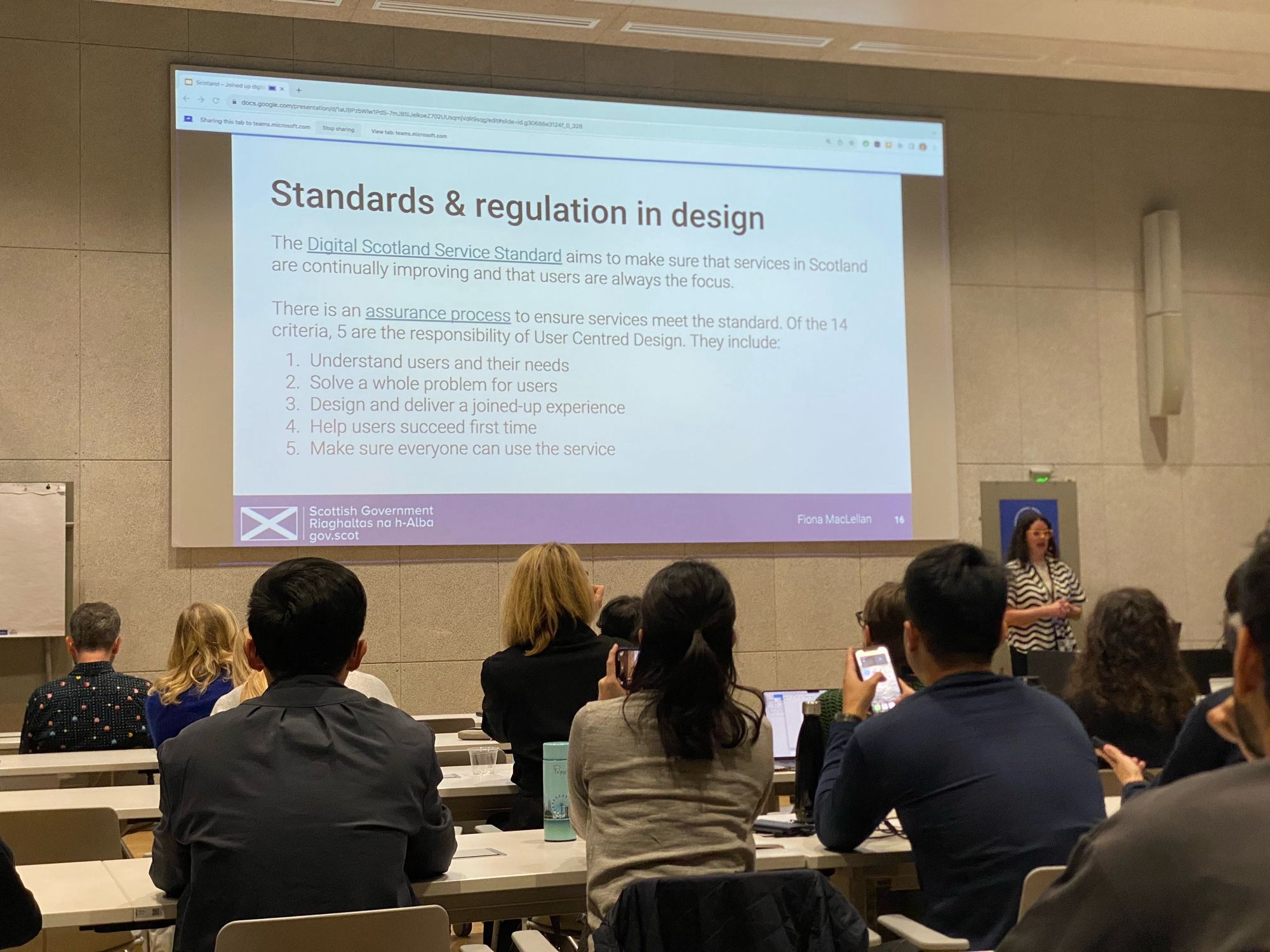 A woman standing at the front of a classroom or conference room, giving a presentation. On the screen behind her is a slide titled "Standards & regulation in design," referring to the Digital Scotland Service Standard. The slide lists five user-centered design responsibilities, including understanding users' needs and ensuring accessibility for all users. The audience is seated at desks, some of them using laptops and mobile devices.