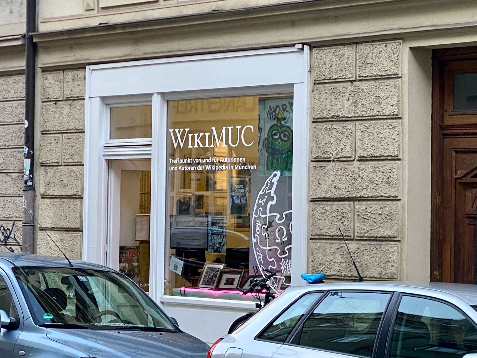 A storefront with a large window displaying the name "WikiMUC." Inside the window, there is a large decal of the Wikipedia logo, a desk with computer monitors, and various framed pictures. Two parked cars are visible in front of the building, and graffiti of a green cartoon character is seen on the wall inside.